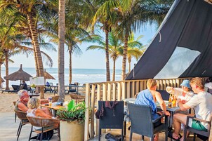 Un área de comedor frente a la playa con mesas y sillas, palmeras y una carpa.