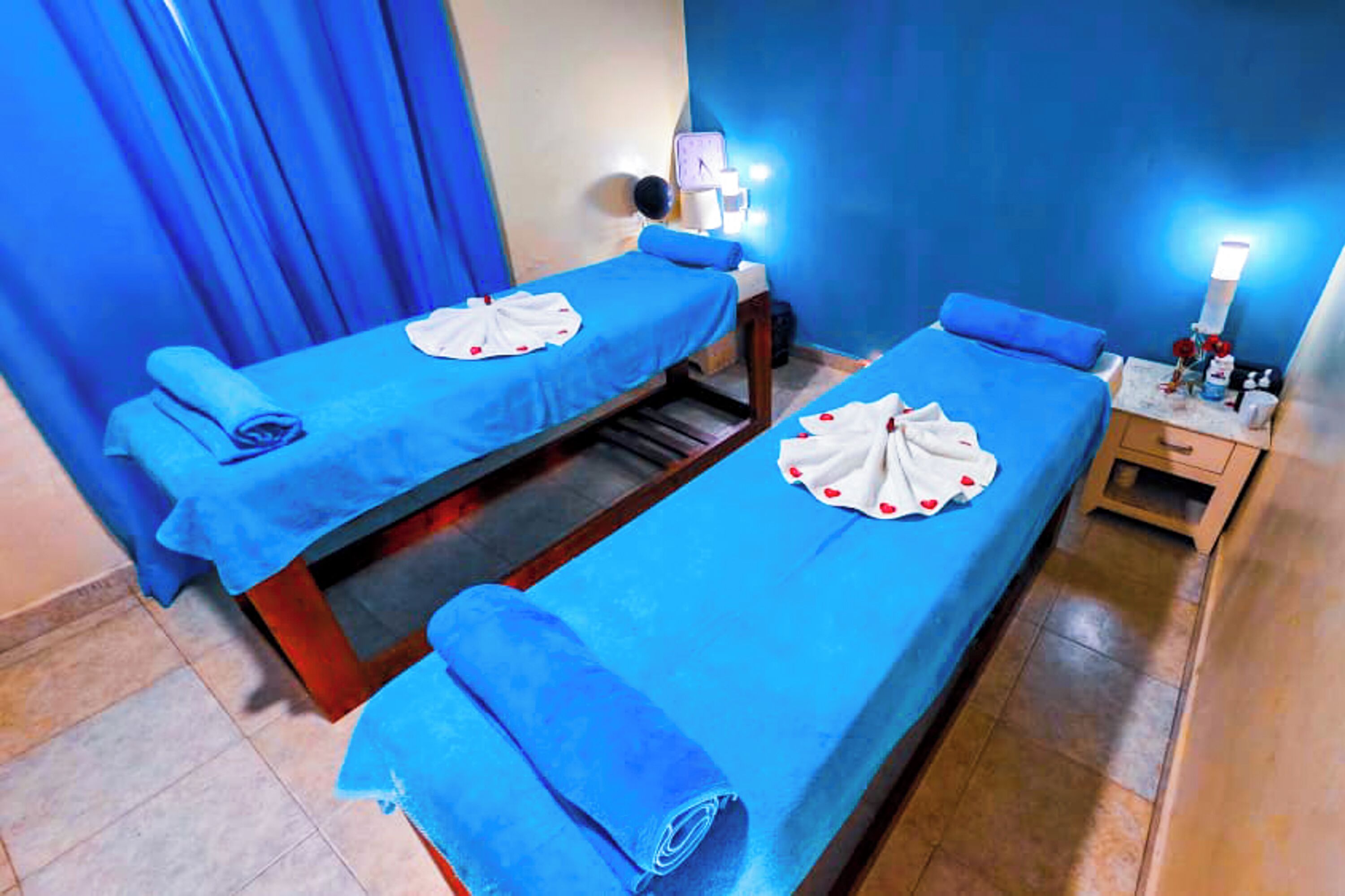 Habitación con dos camas, cada una con cubrecama azul y toalla blanca con diseños de corazones rojos. Hay lámparas de noche y una mesita con artículos de aseo.