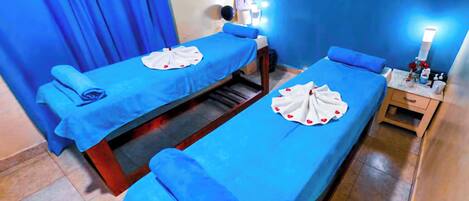 A room with two beds, each with a blue bedspread and a white towel with red heart designs. There are bedside lamps and a small table with toiletries.