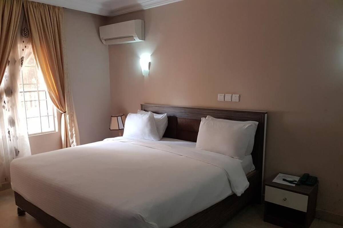 A neatly made bed with white linens, a wooden headboard, a bedside table with a lamp, and an air conditioning unit on the wall.