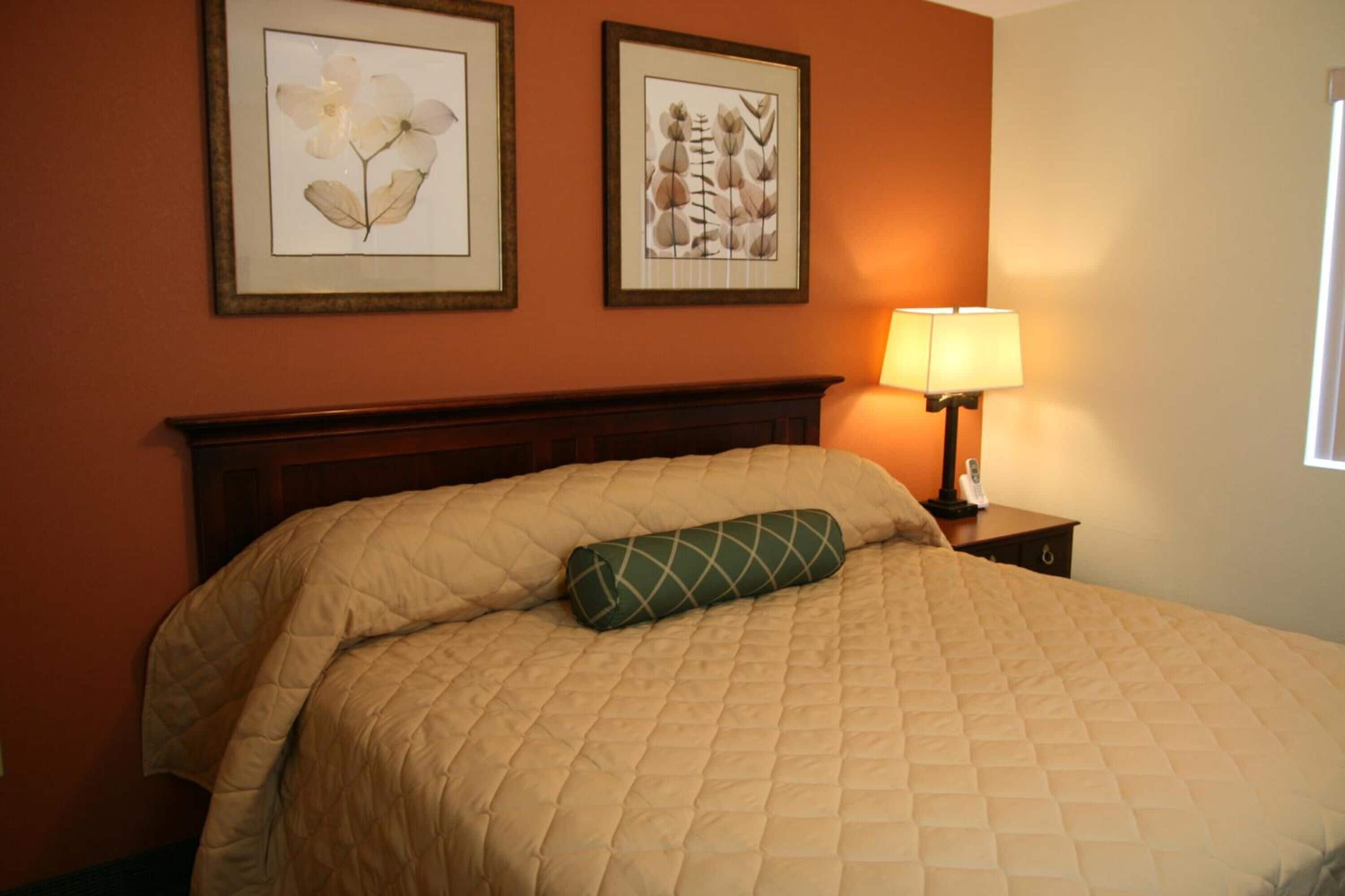 A neatly made bed with a quilted blanket and a decorative pillow, a bedside lamp, and two framed botanical prints on the wall.