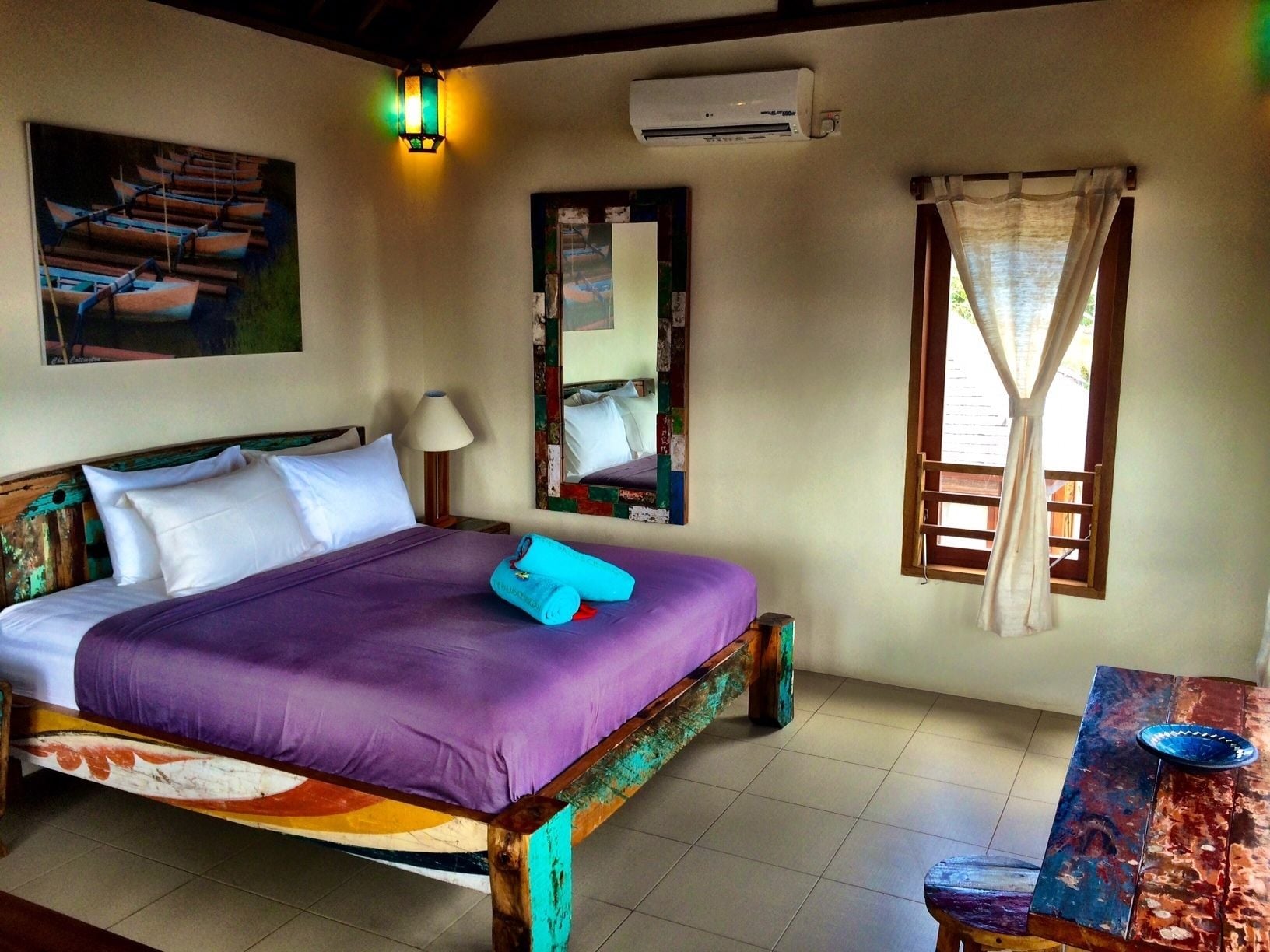 Una habitación con cama, mesita de noche, lámpara, ventana con cortinas, un cuadro y un equipo de aire acondicionado.