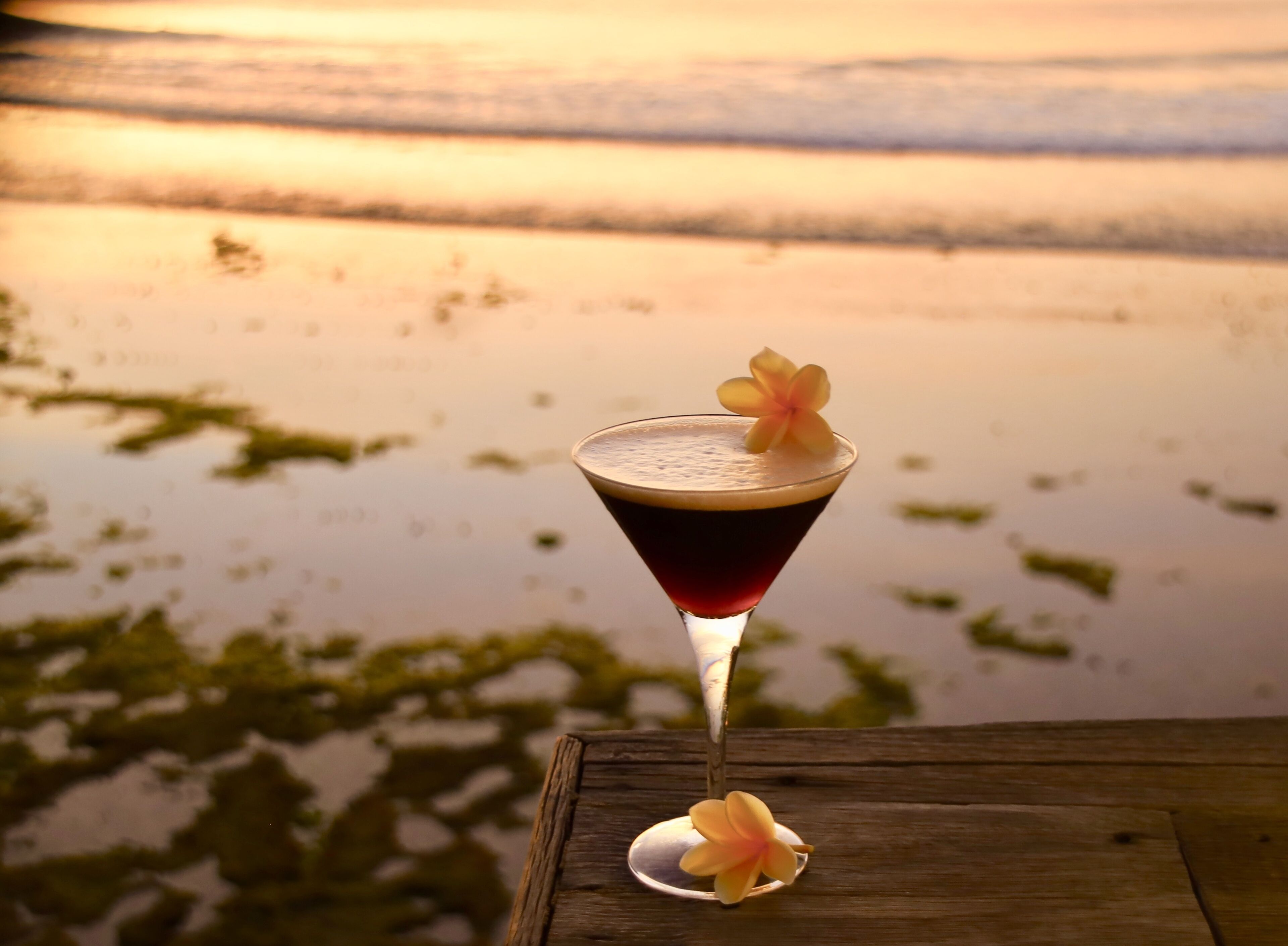 A cocktail on a wooden table with a view of a calm body of water and a sunset in the background.