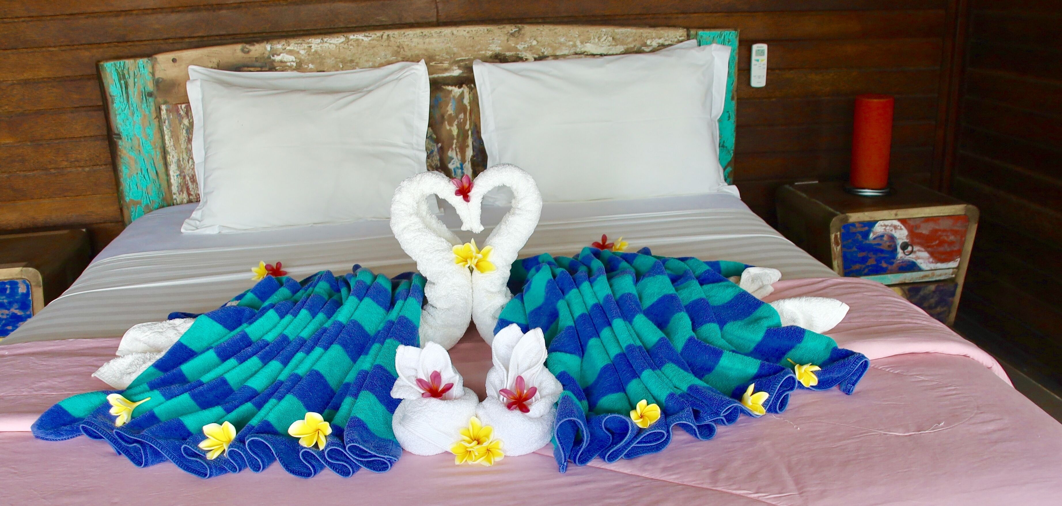 A bed with a blue and white checkered blanket, white pillows, and a heart-shaped towel decoration.