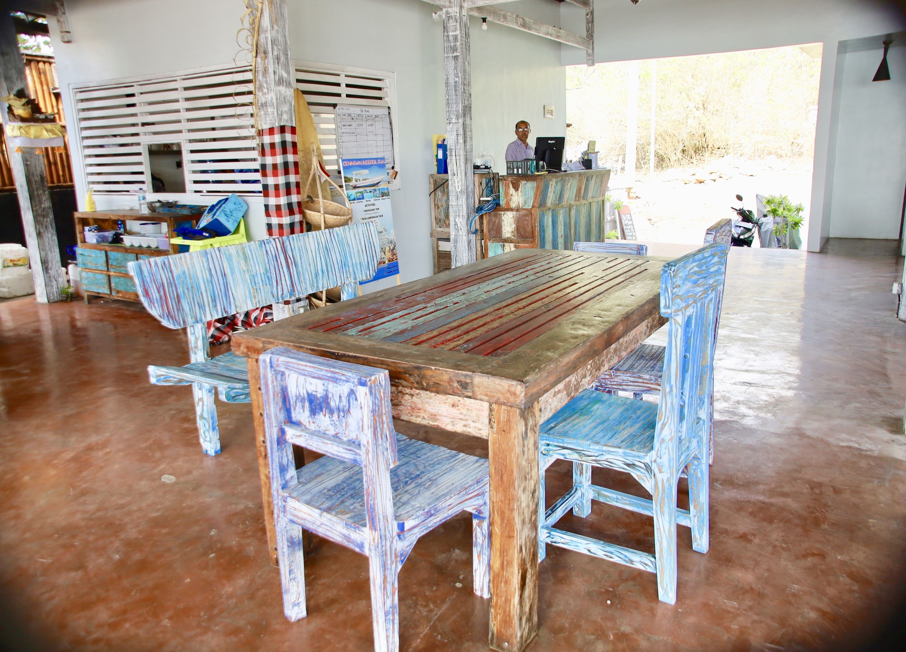 A rustic wooden table with benches, a window with blinds, and a person working at a desk.