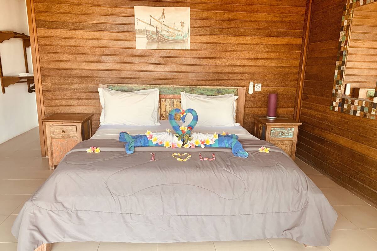 A neatly made bed with a gray bedspread, two pillows, and a decorative heart-shaped arrangement made from towels and flowers.
