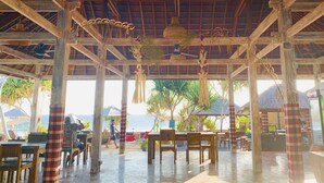 An open-air dining area with wooden tables and chairs, a thatched roof, and views of the sea. - The Palms Ceningan Hotel (Ceningan Island)