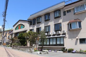A multi-story hotel with a modern facade, large windows, and a small garden area with various plants.