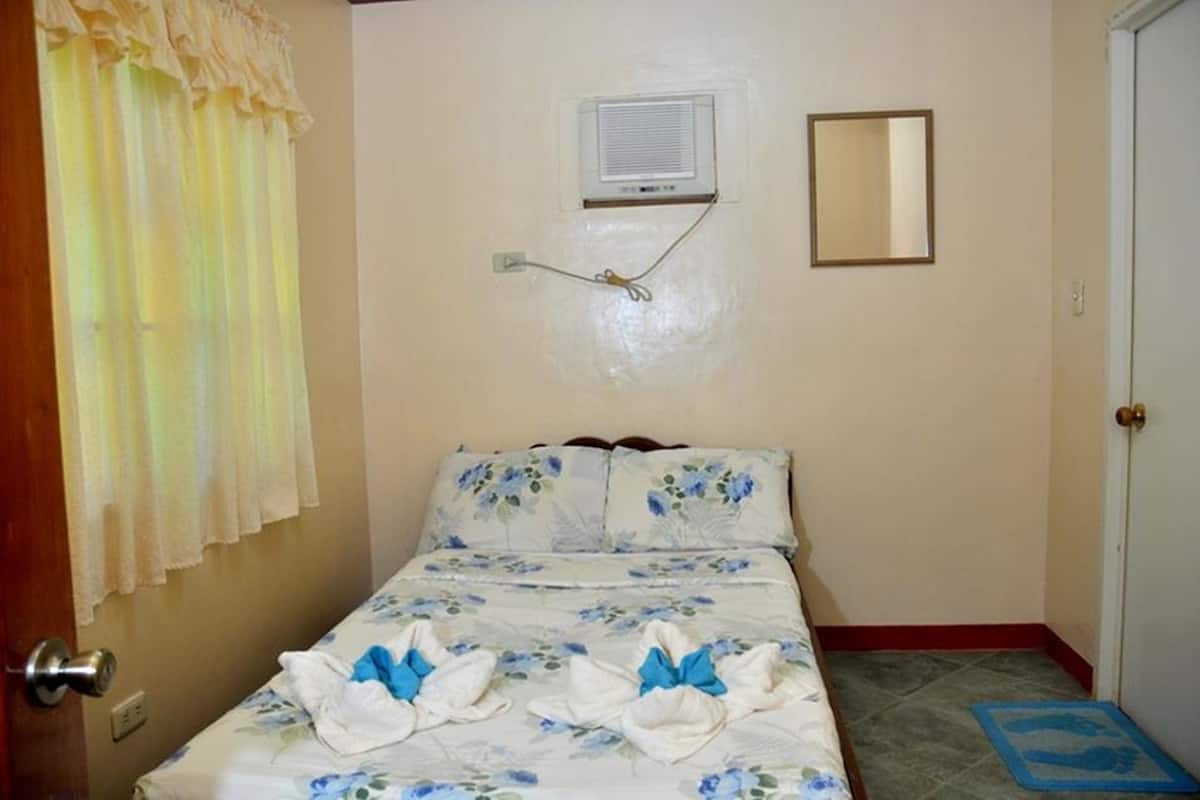 Un dormitorio con cama, ventana con cortinas, unidad de aire acondicionado, espejo y puerta.