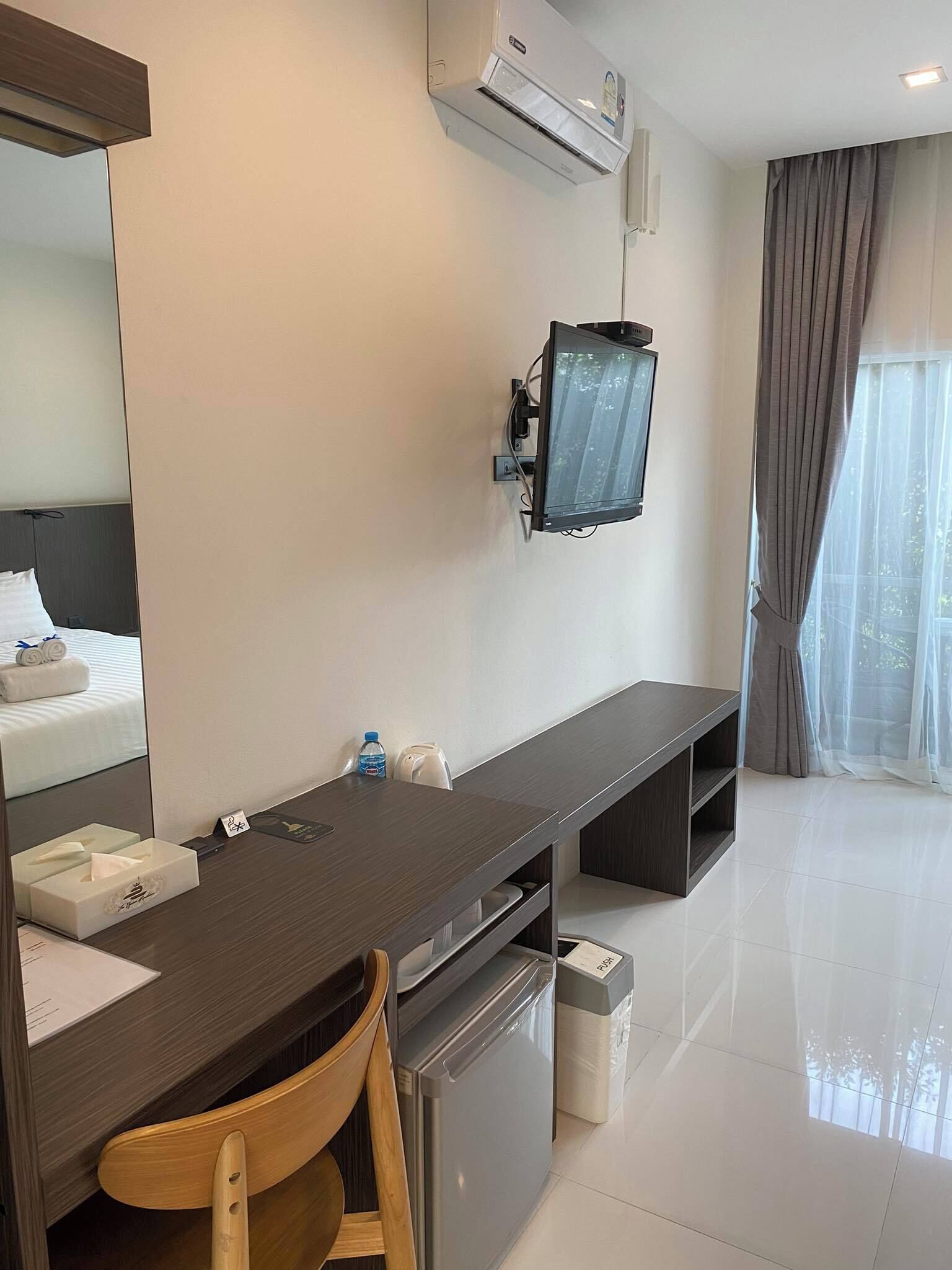 A hotel room with a long dark wooden desk, a mirror, a wall-mounted TV, a small refrigerator, a trash bin, and a window with curtains.