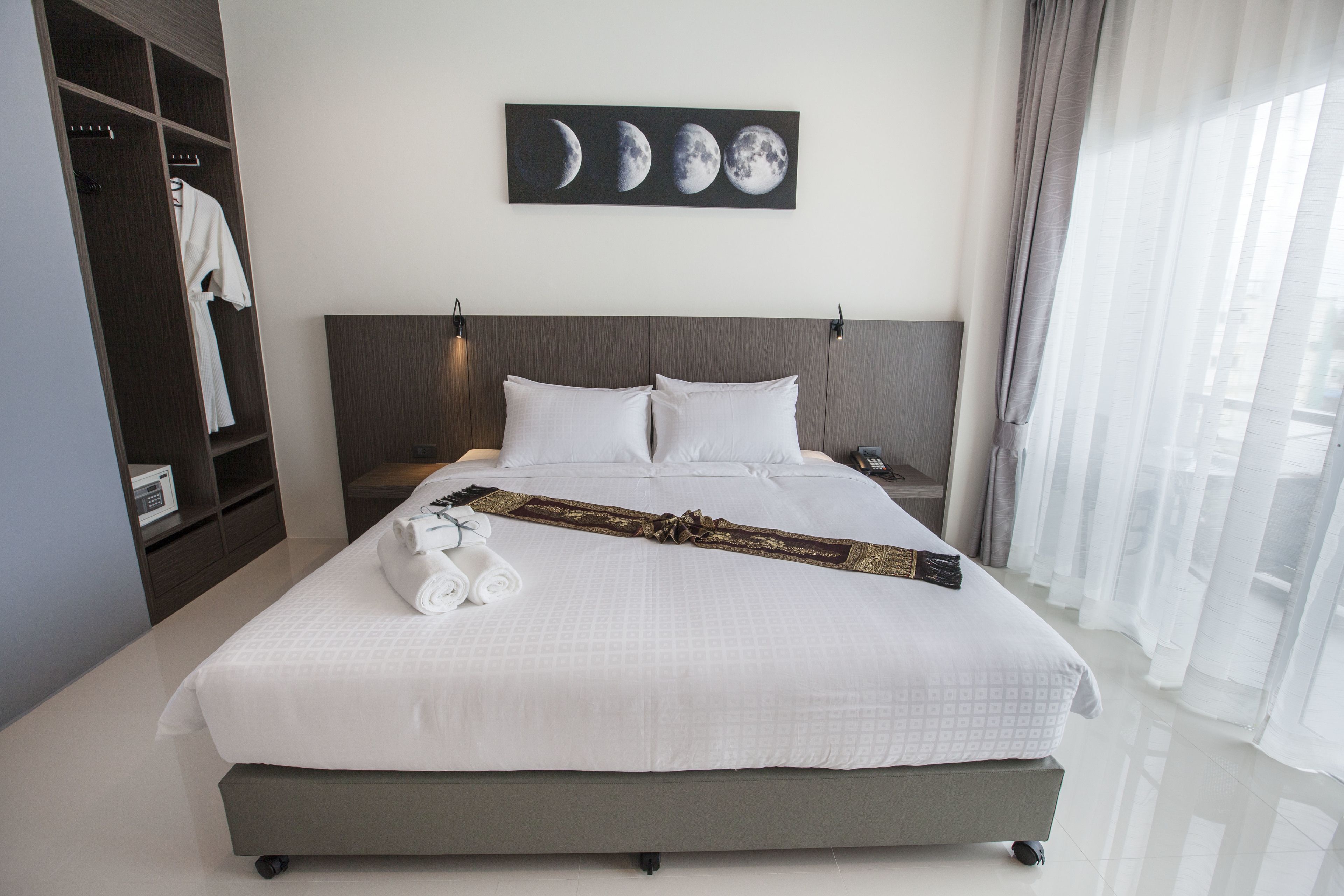 A neatly made bed with white linens and a decorative headboard, a wardrobe with a hanging robe, and a wall-mounted artwork depicting the moon phases.
