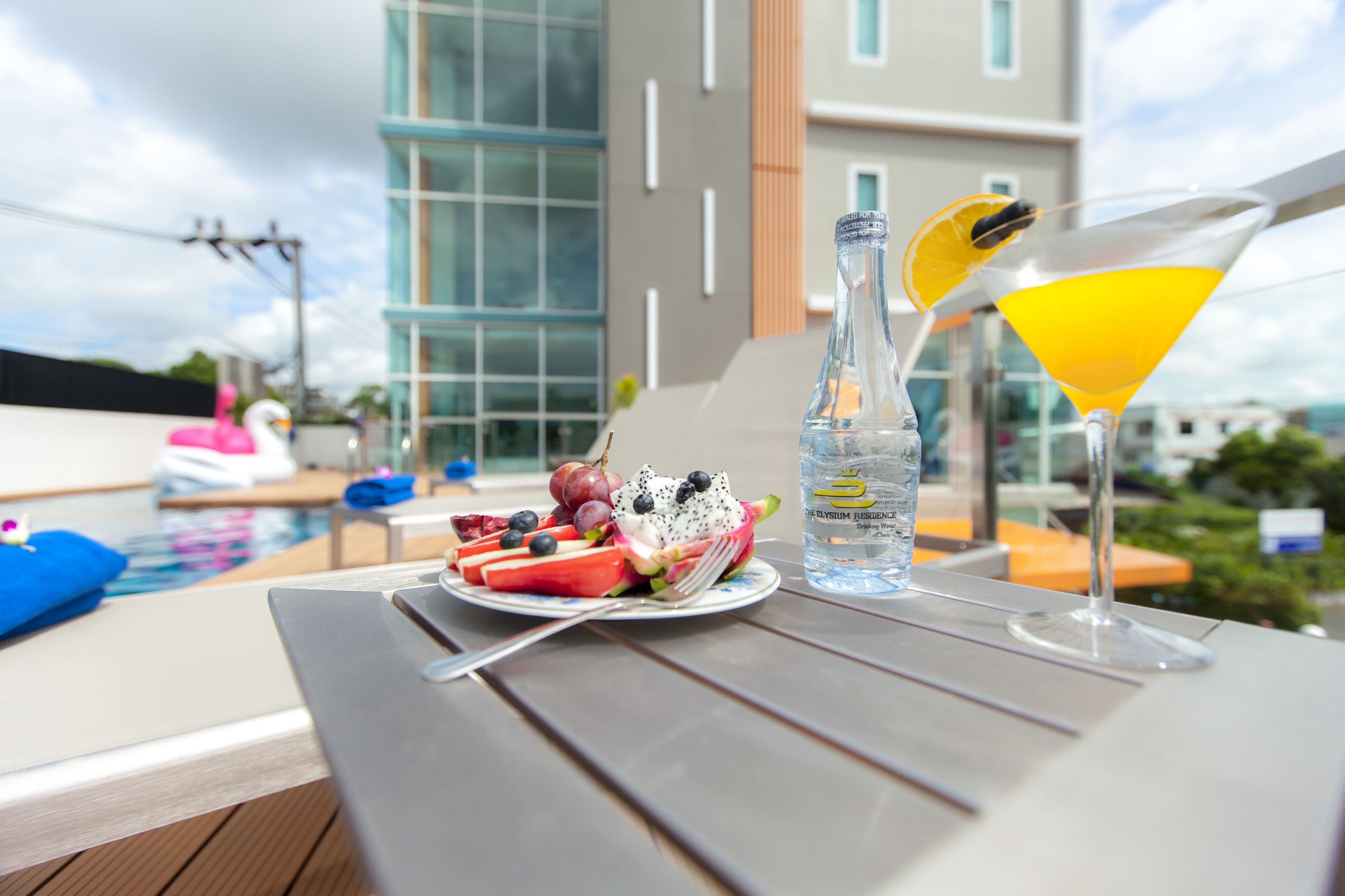 Een tafel met een fruitschaal, een waterfles en een martini-glas op een balkon met uitzicht op een zwembad en moderne gebouwen.