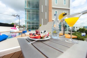 Una mesa con un plato de fruta, una botella de agua y una copa de martini en un balcón con vista a una piscina y edificios modernos.