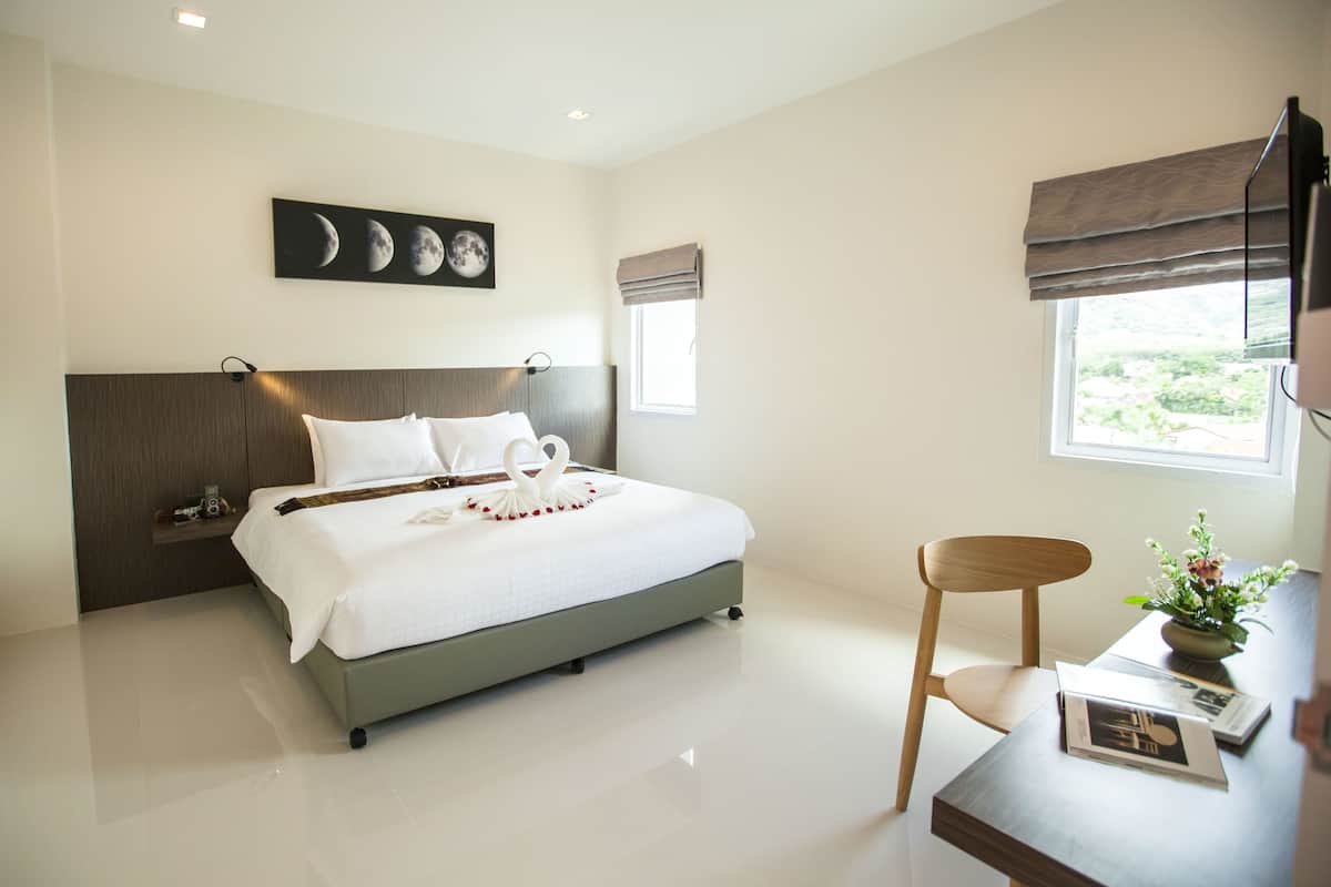 A modern hotel room with a large bed, a wooden headboard, a bedside table with a vase of flowers, and a window with a view of greenery.