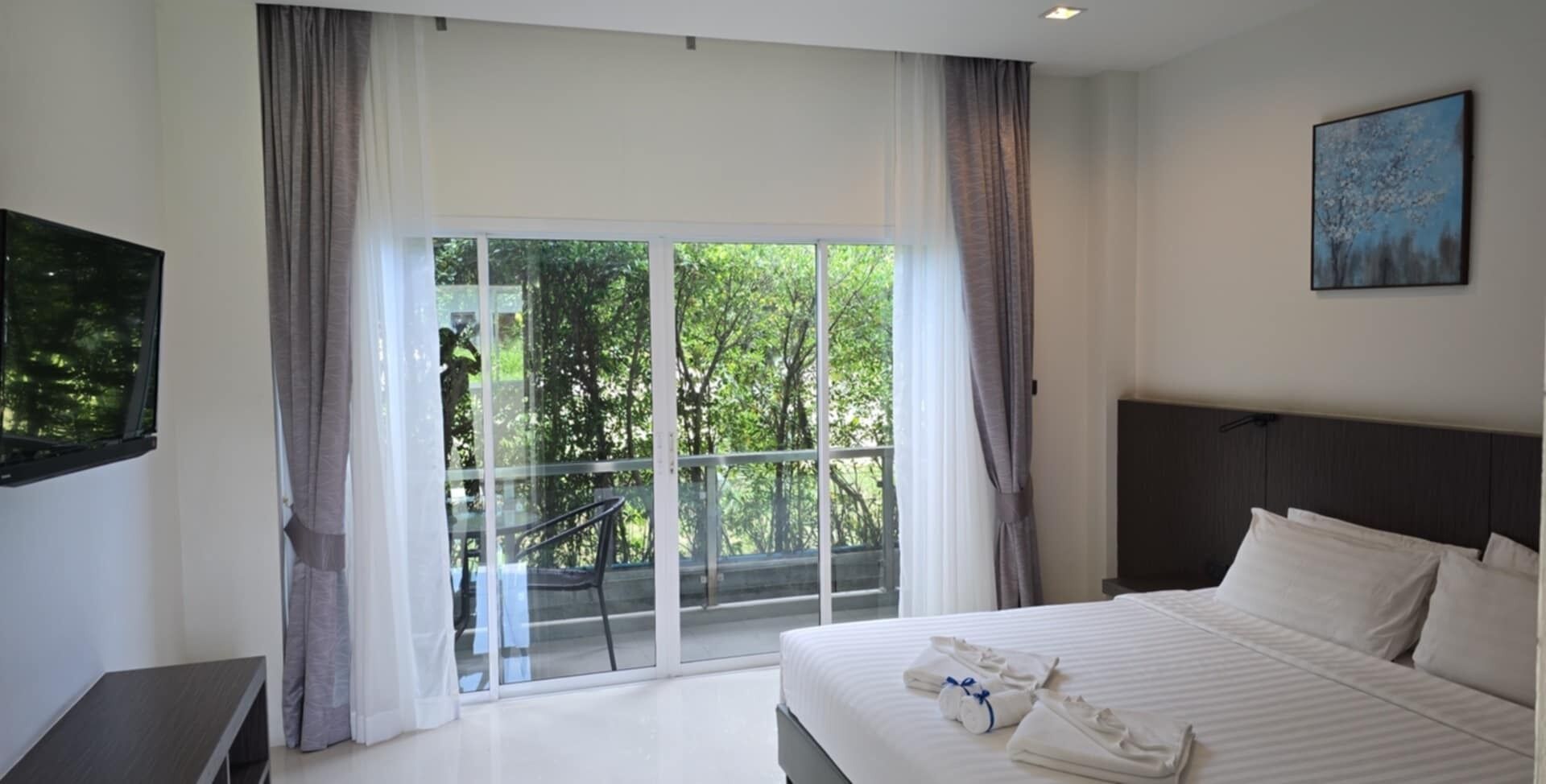 A hotel room with a large bed, a flat-screen TV, a balcony with a view of trees, and a painting on the wall.