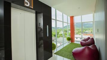 Een moderne hotel lobby met een groot raam met uitzicht op de stad en heuvels. Er zijn twee rode relaxfauteuils en een kleine groene plant op een groen tapijt.