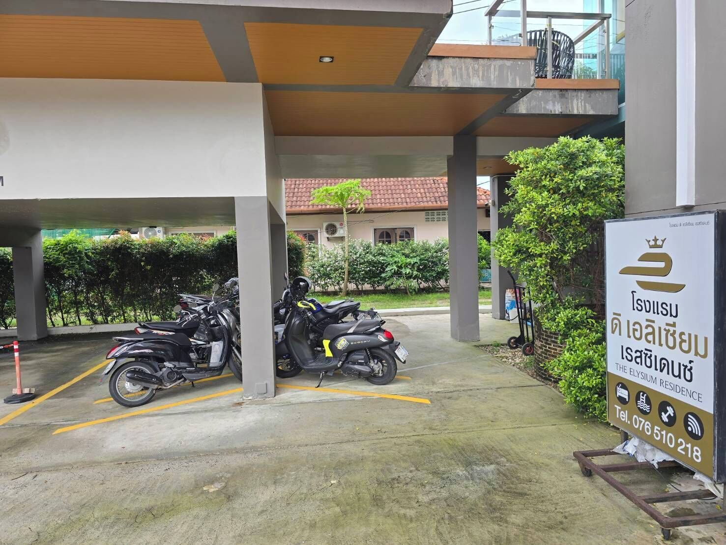 A parking area with motorcycles, a signboard for The Elysium Residence, and a building with a tiled roof in the background.
