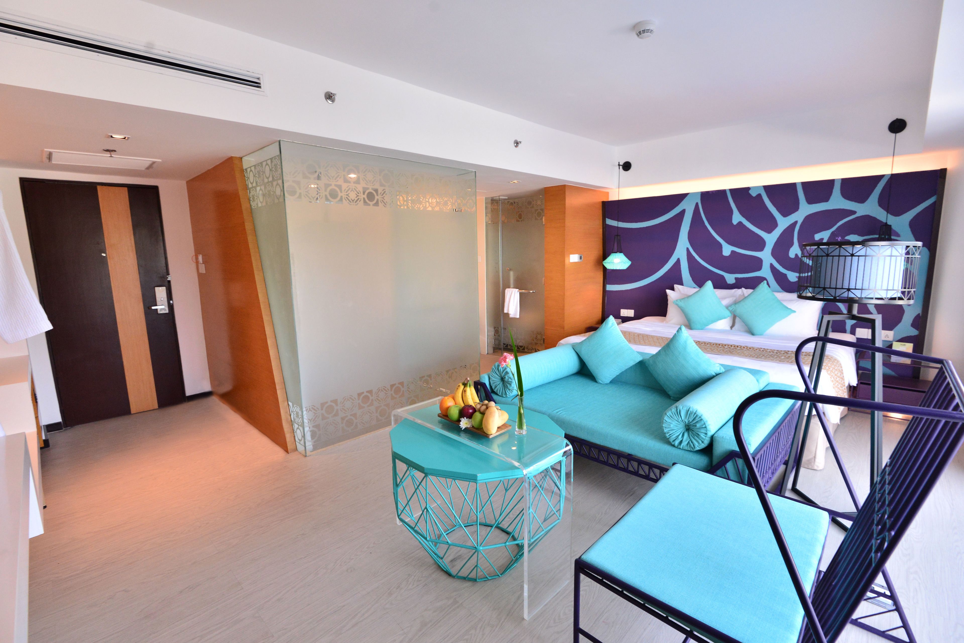 Une chambre d’hôtel moderne avec une cloison en verre, un canapé turquoise, une petite table avec des fruits et un lit avec une literie bleue.