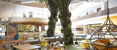 Un espacio interior moderno con un árbol central, zonas de estar y un bar.