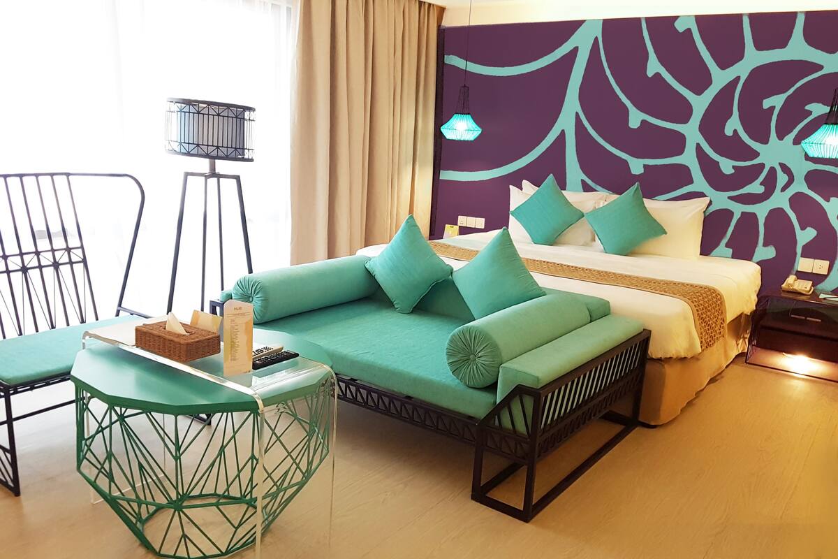 A modern hotel room with a green sofa, a bed with white bedding, a green accent chair, and a geometric-patterned wall.