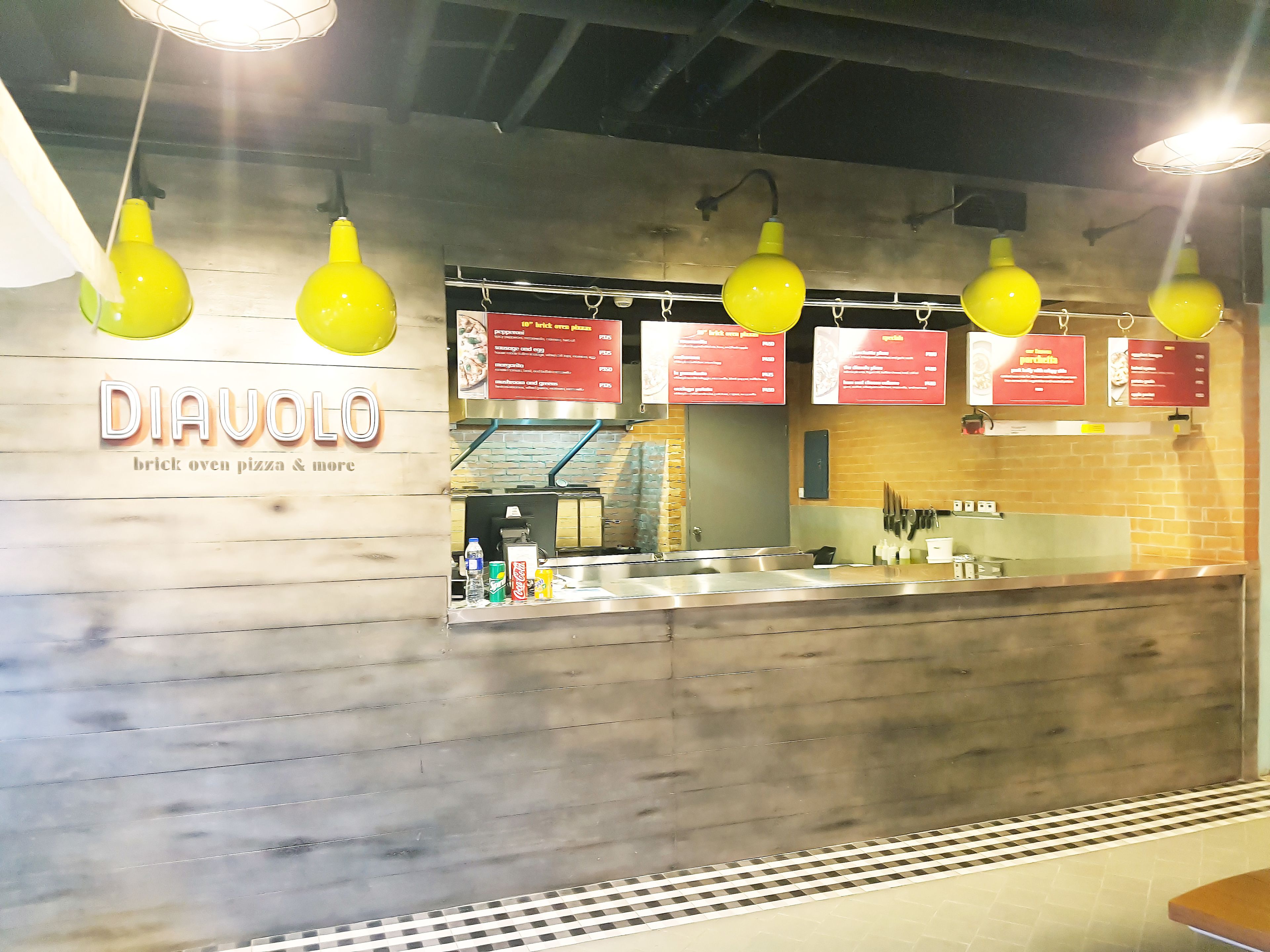 Intérieur d’un restaurant servant des pizzas cuites au four à briques, avec des lampes jaunes suspendues, un tableau affichant le menu et un comptoir où sont exposés divers produits.