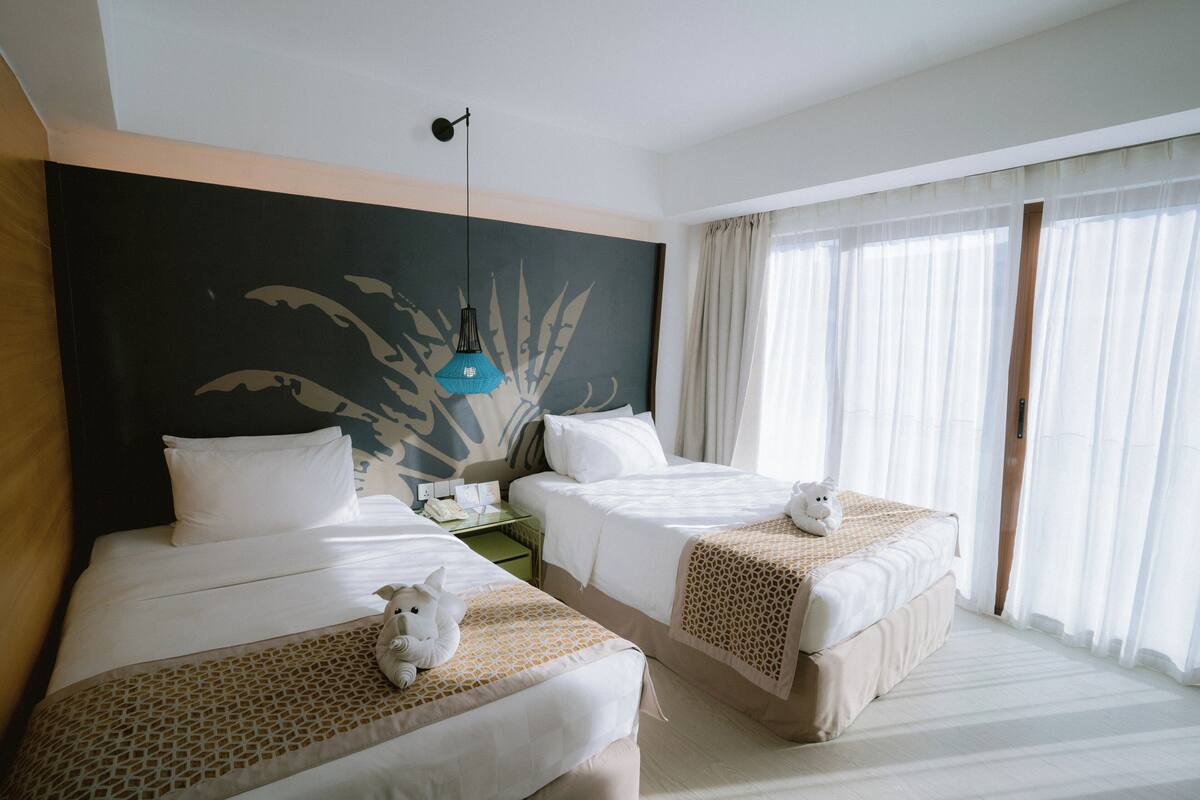 A hotel room with two beds, a large wall mural, a desk with a lamp, and a view of the outdoors through sheer curtains.