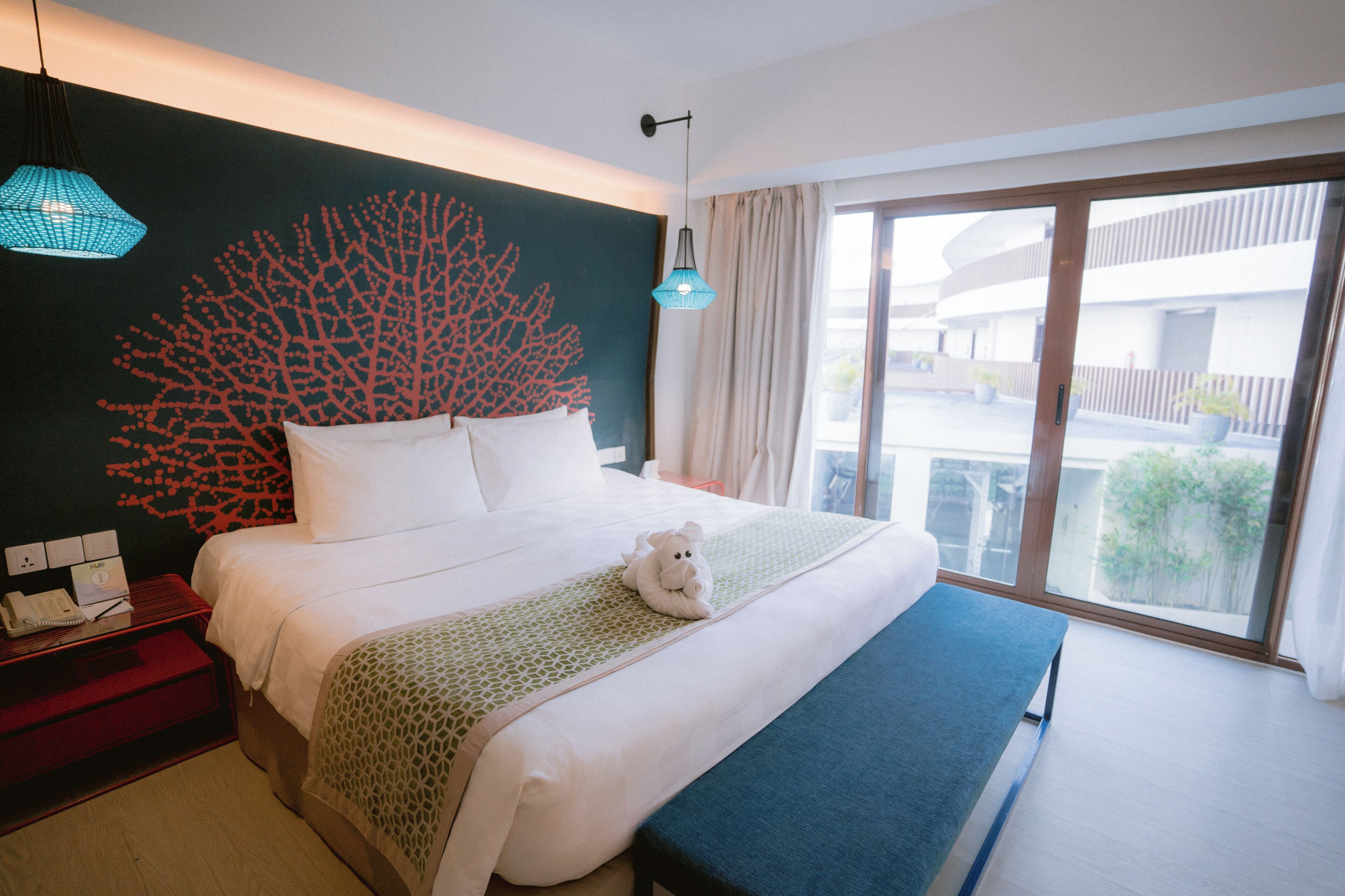 Habitación de hotel con una cama grande, una mesita de noche con teléfono y bloc de notas, una obra de arte en la pared y vistas al balcón exterior.