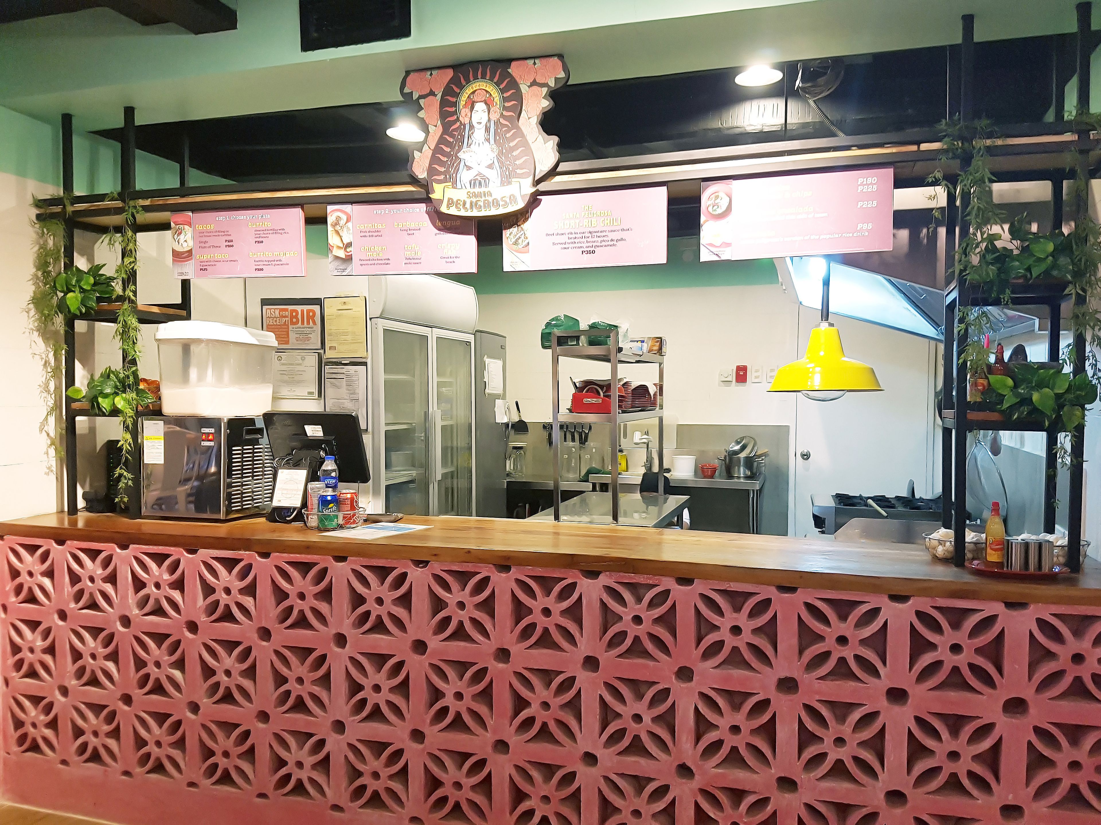 A restaurant counter with a pink decorative panel, a menu board, and a yellow pendant light.
