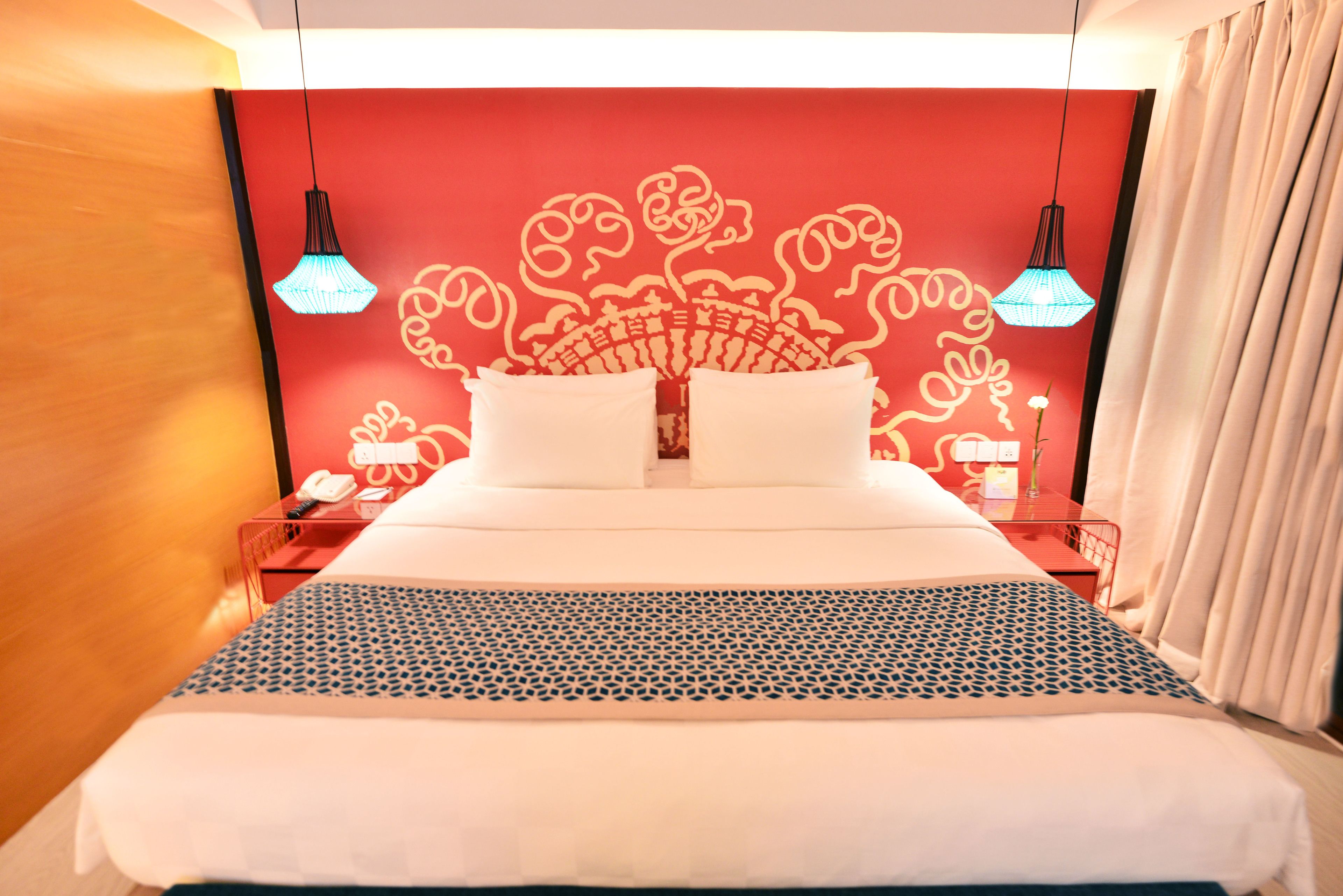 A hotel room with a large bed, red wall decor, two bedside lamps, and a nightstand with a phone and a vase.