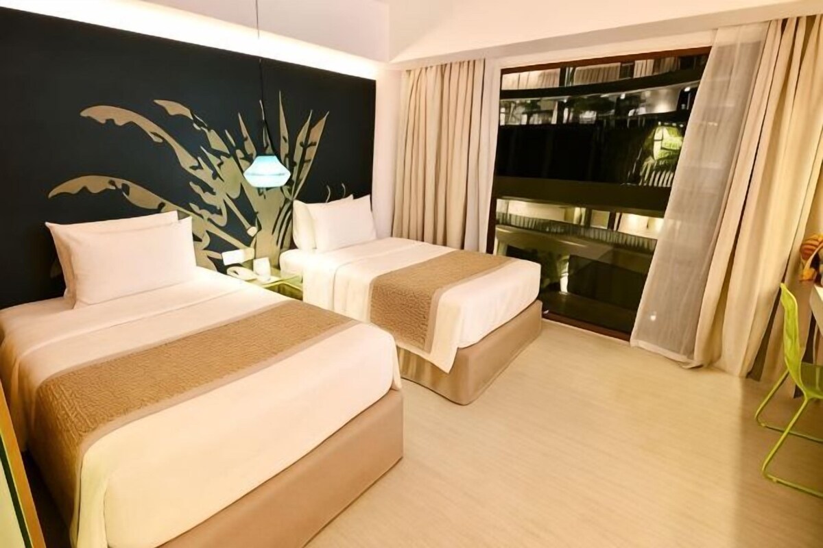 Habitación de hotel con dos camas, una mesita de noche, una planta y un balcón con cortinas.