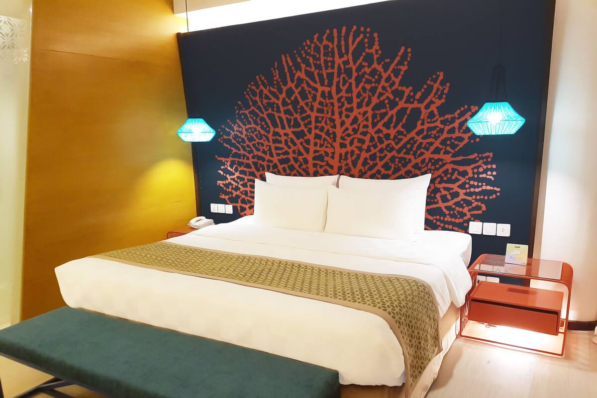 A neatly made bed with a patterned bedspread, a bedside table with a lamp, and a wall with a coral tree mural.