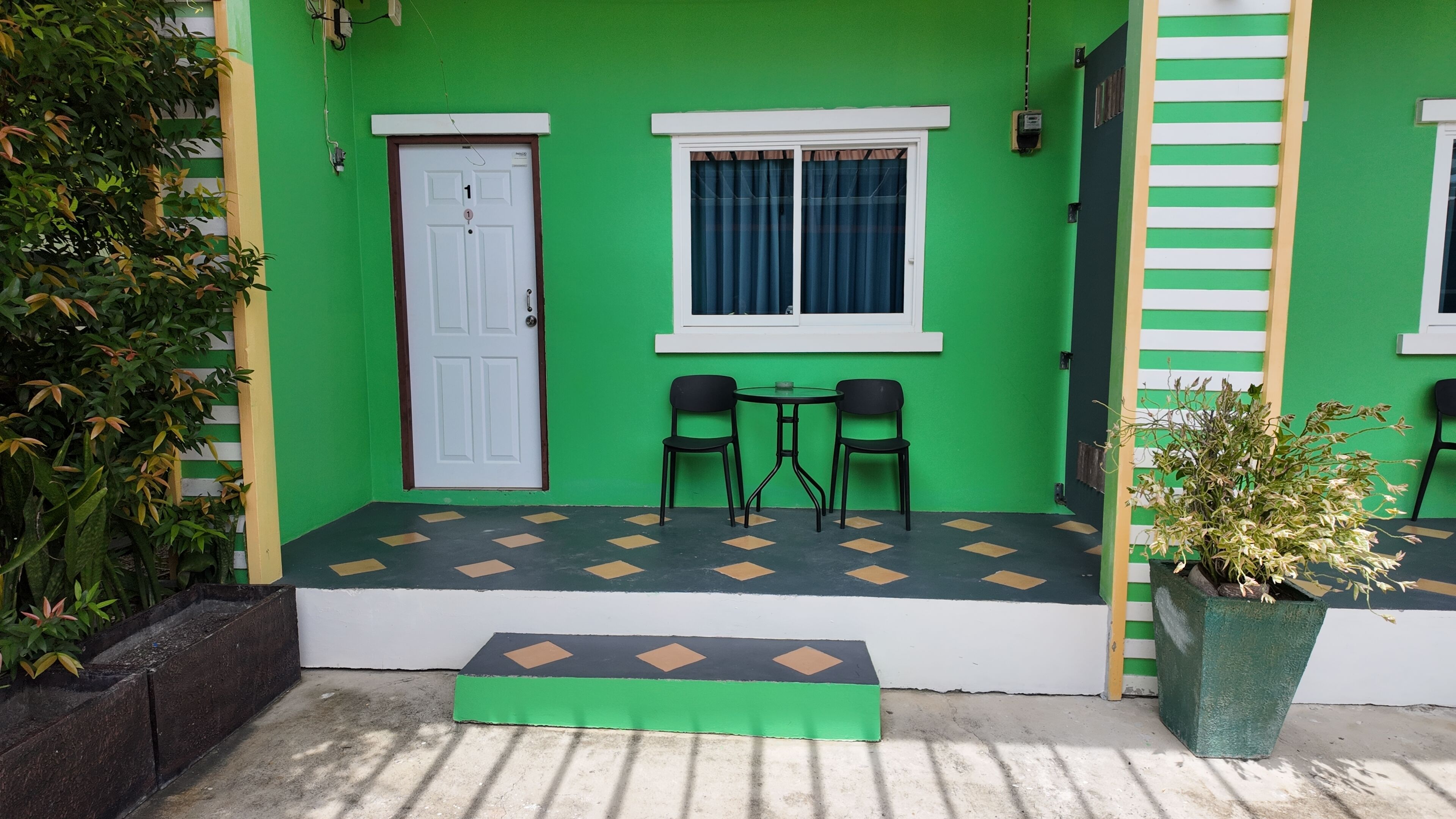Un edificio verde con una puerta blanca, una ventana con cortinas y dos sillas negras afuera.