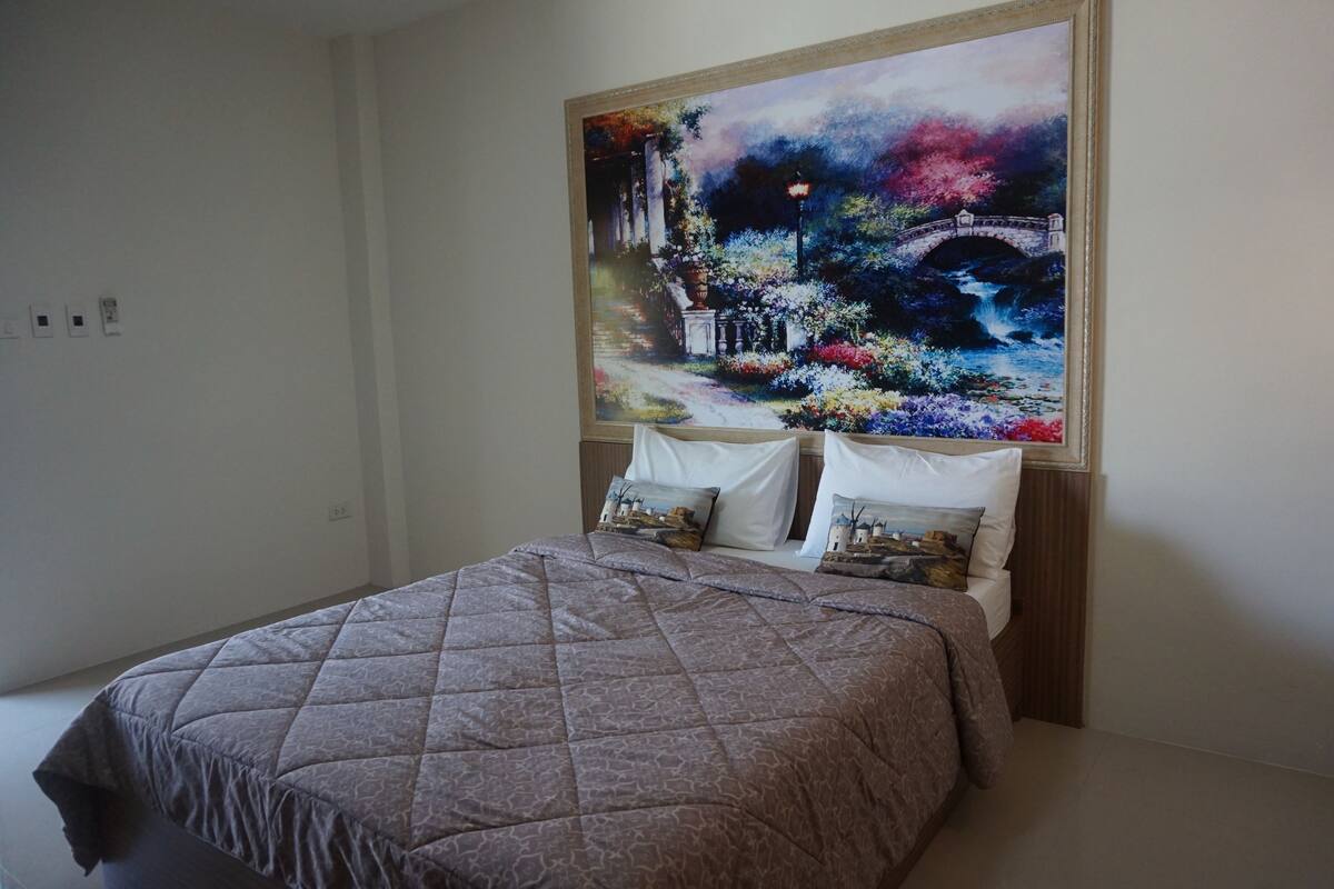 A bedroom with a bed, a large painting of a garden scene, and a night light.