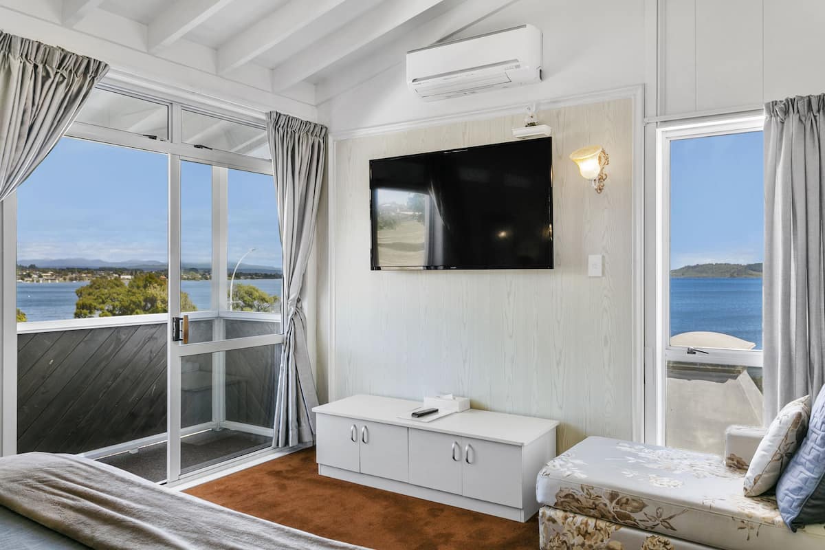 Une chambre avec une grande fenêtre, un lit, une commode blanche et un téléviseur à écran plat fixé au mur.