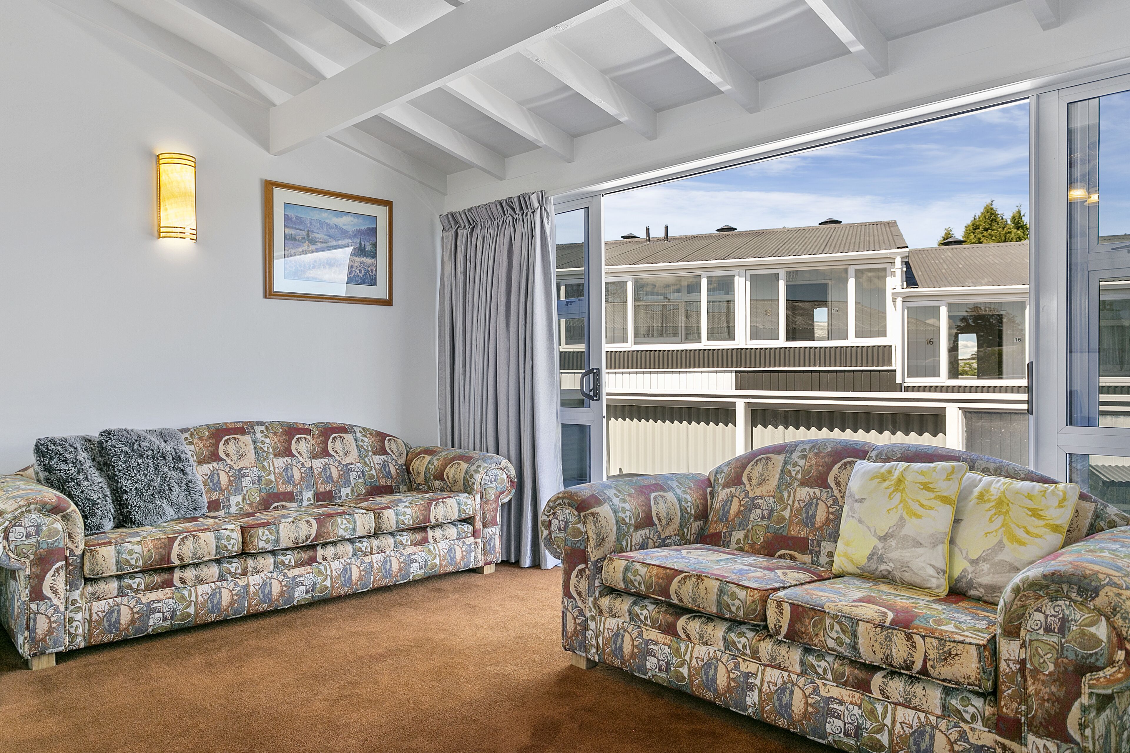 A modern living room with two patterned sofas, a large window with a view of a house, and a framed picture on the wall.