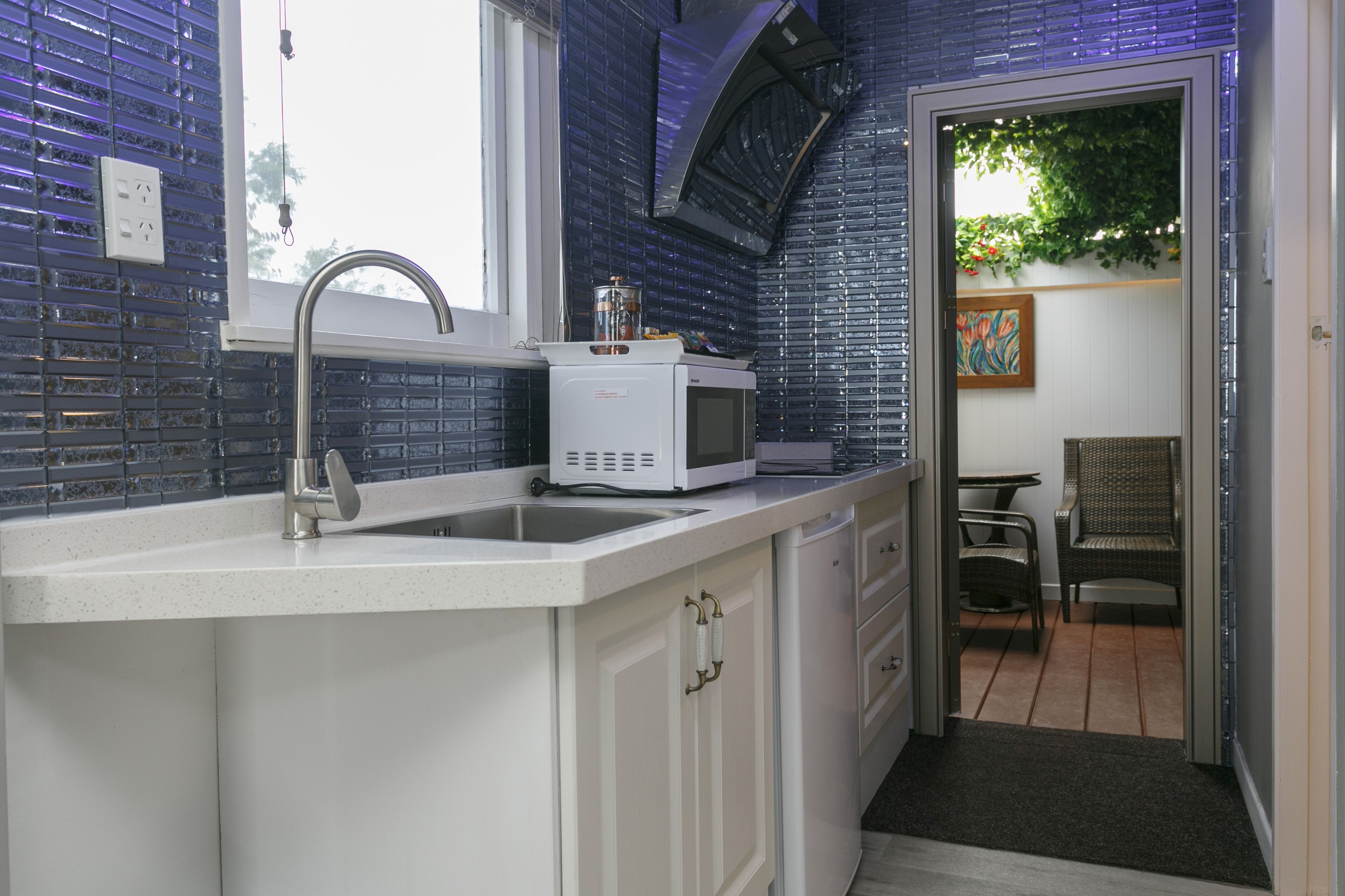 A kitchen with a white countertop, a sink, and a microwave. There is a window above the sink and a door leading to a dining area with a chair and table.