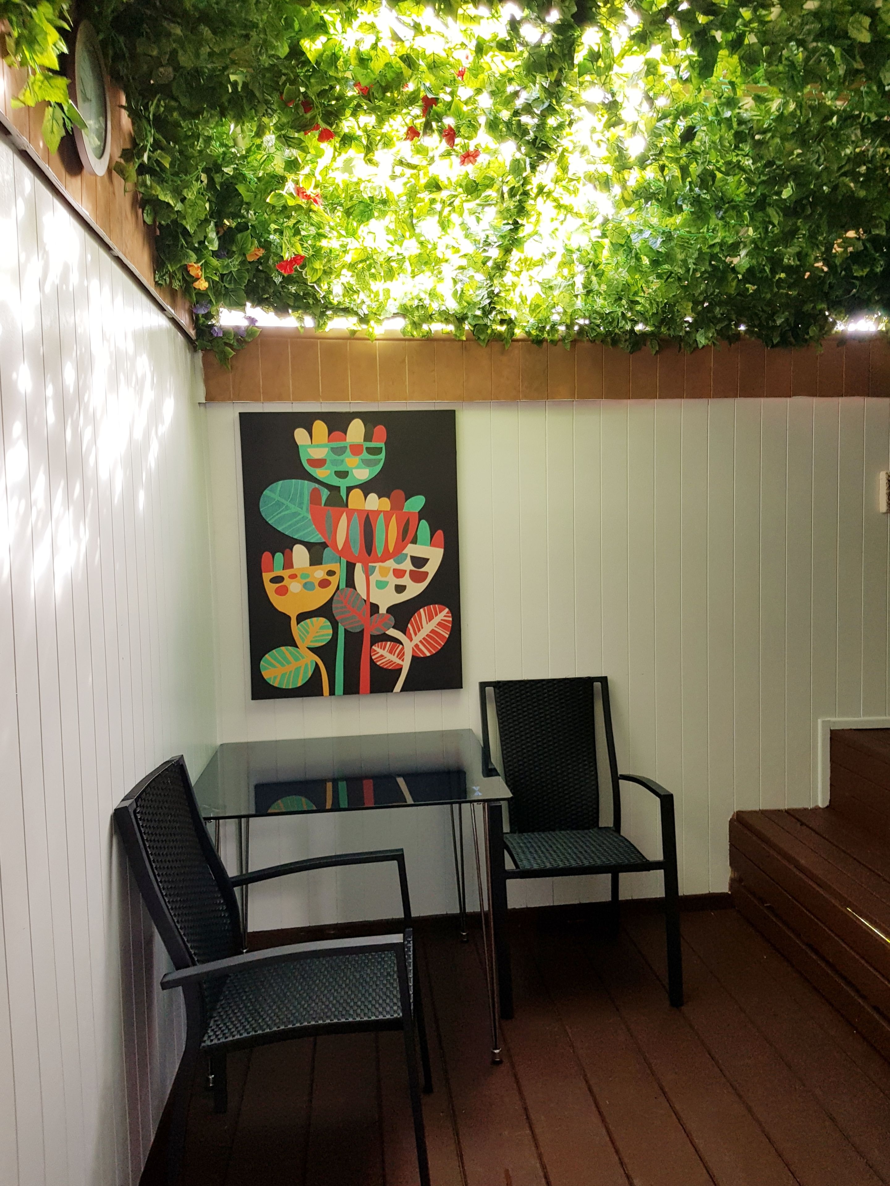 Un espace patio avec une table en verre et deux chaises, une peinture abstraite aux couleurs vives accrochée au mur et un banc en bois.