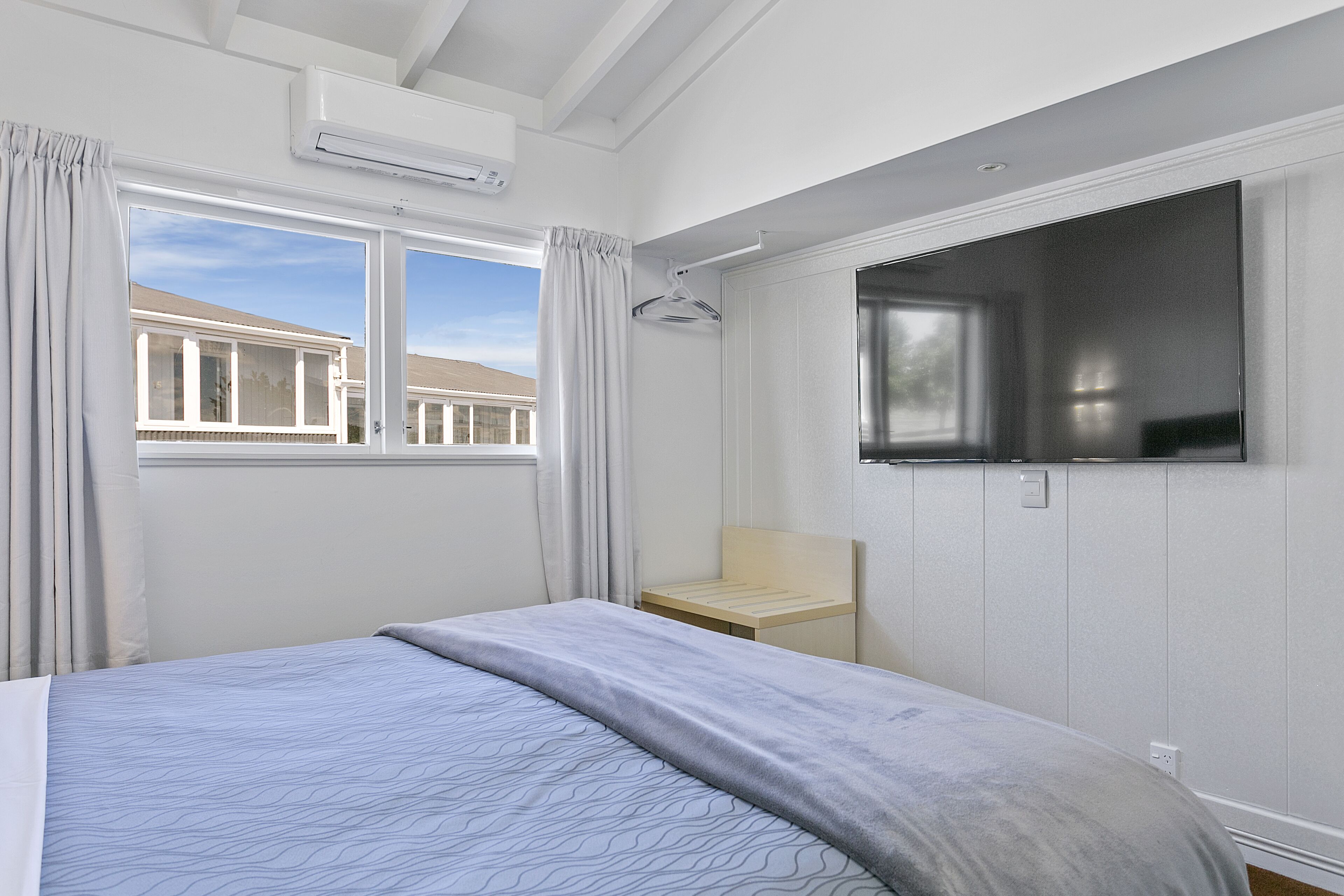 A bedroom with a bed, a TV mounted on the wall, a window with curtains, and a small wooden table.