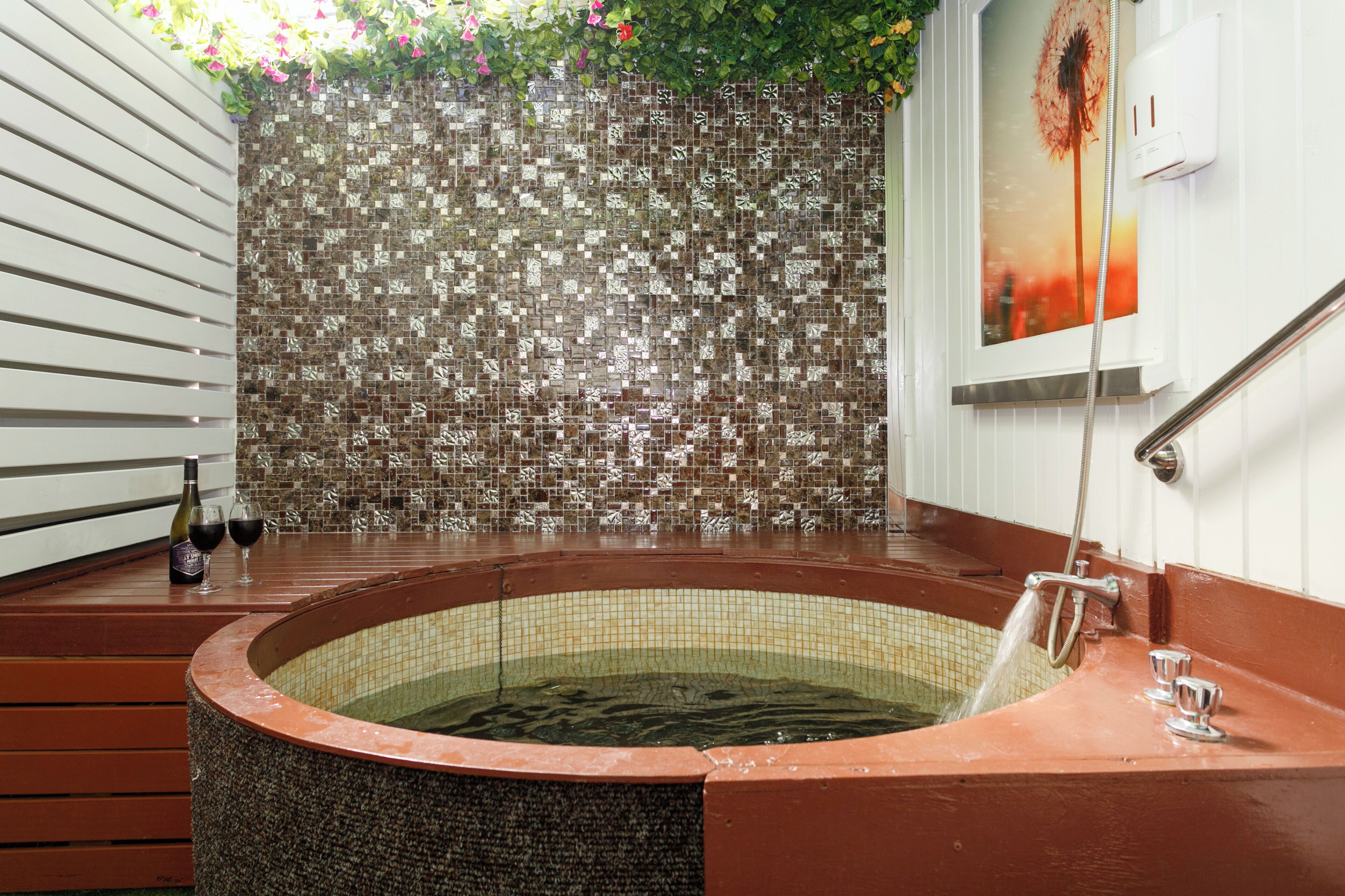 A hot tub with a tiled wall behind it, a bottle of wine, and two wine glasses on the side.