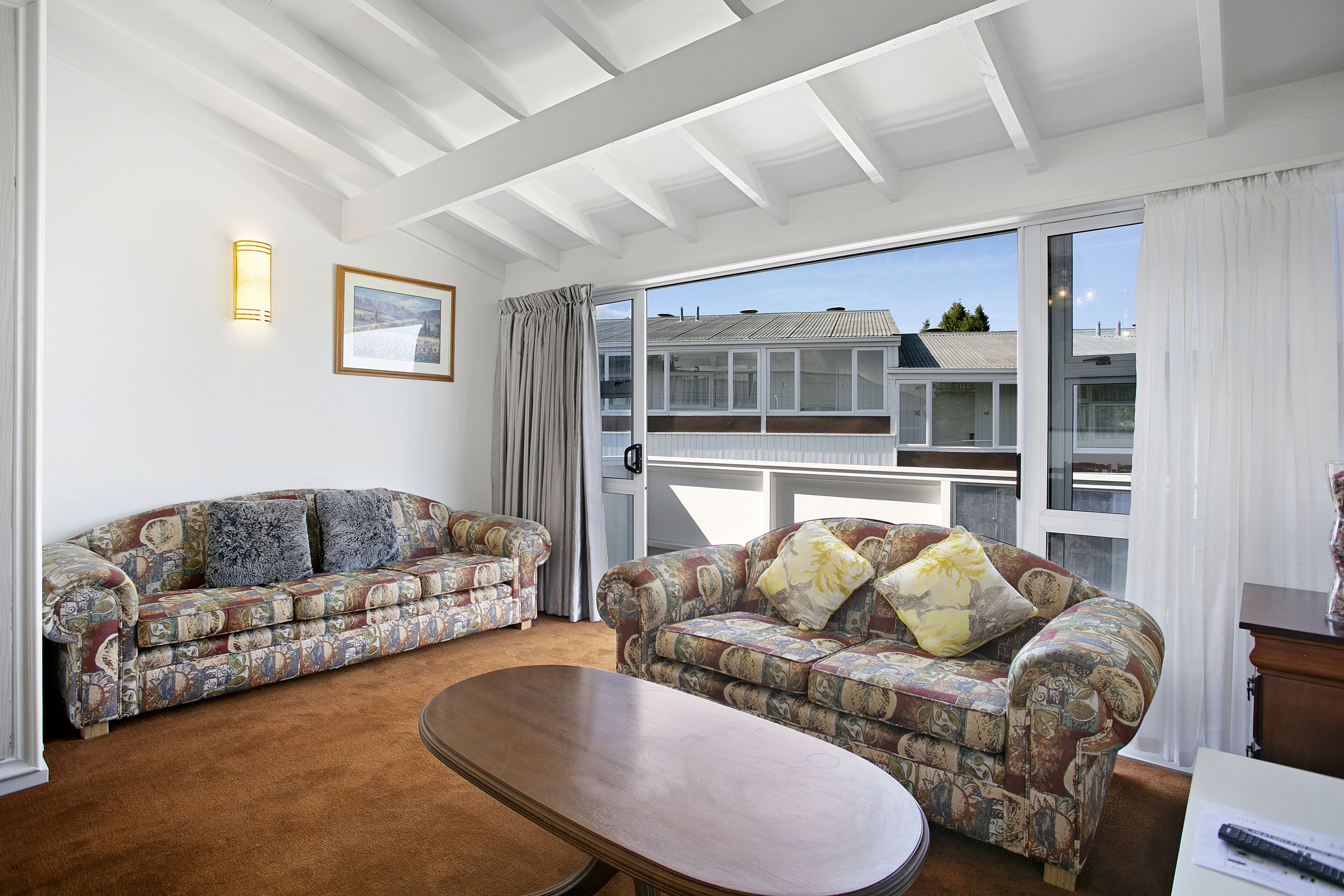 Uma sala de estar com dois sofás estampados, uma mesa de centro de madeira e uma grande janela com vista para edifícios.