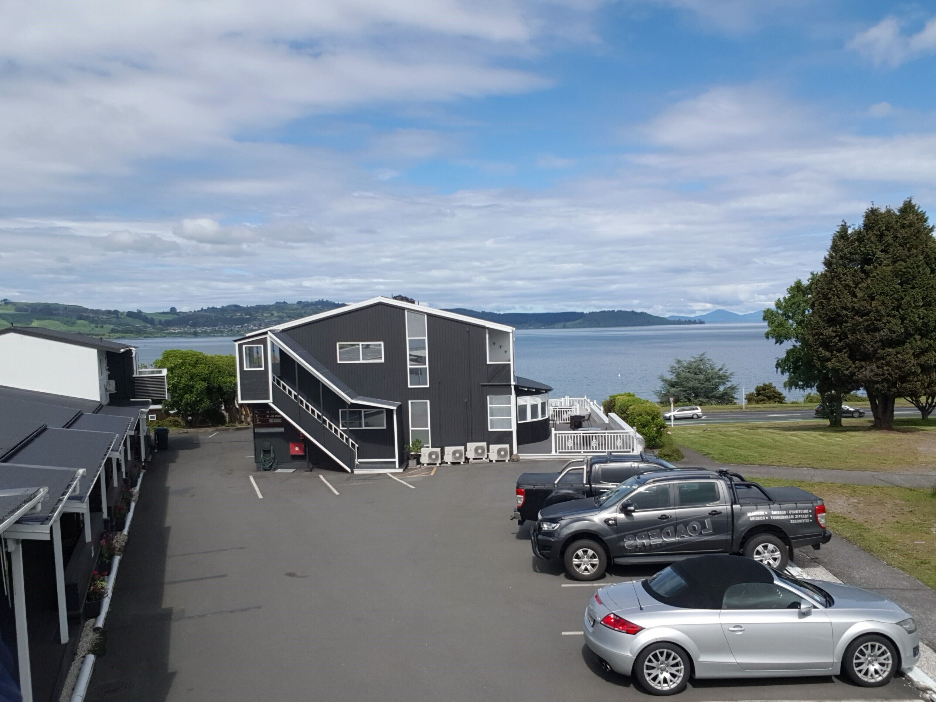A parking lot with a building that has a staircase leading to a deck. There are cars parked in the lot, and a view of a body of water and hills in the background.