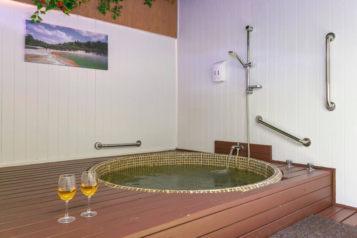 Une salle de spa équipée d’un jacuzzi, de barres d’appui, d’un tableau représentant une plage et de deux verres de vin.