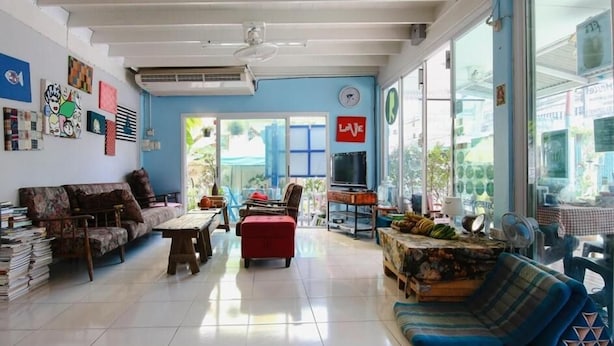 Amplia sala de estar con sofá, un taburete rojo, televisor y mesa de comedor. Grandes ventanas ofrecen luz natural y vistas al exterior.