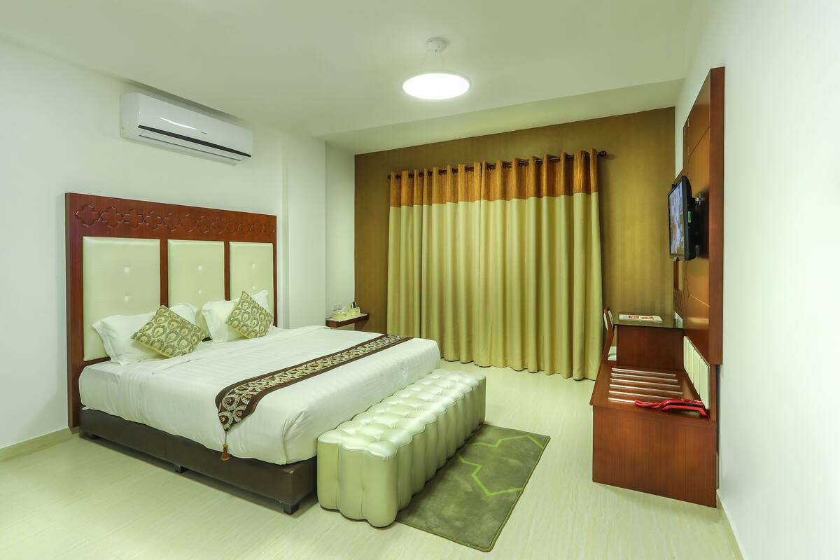 A hotel room with a bed, bedside tables, an air conditioning unit, a television, and a window with curtains.