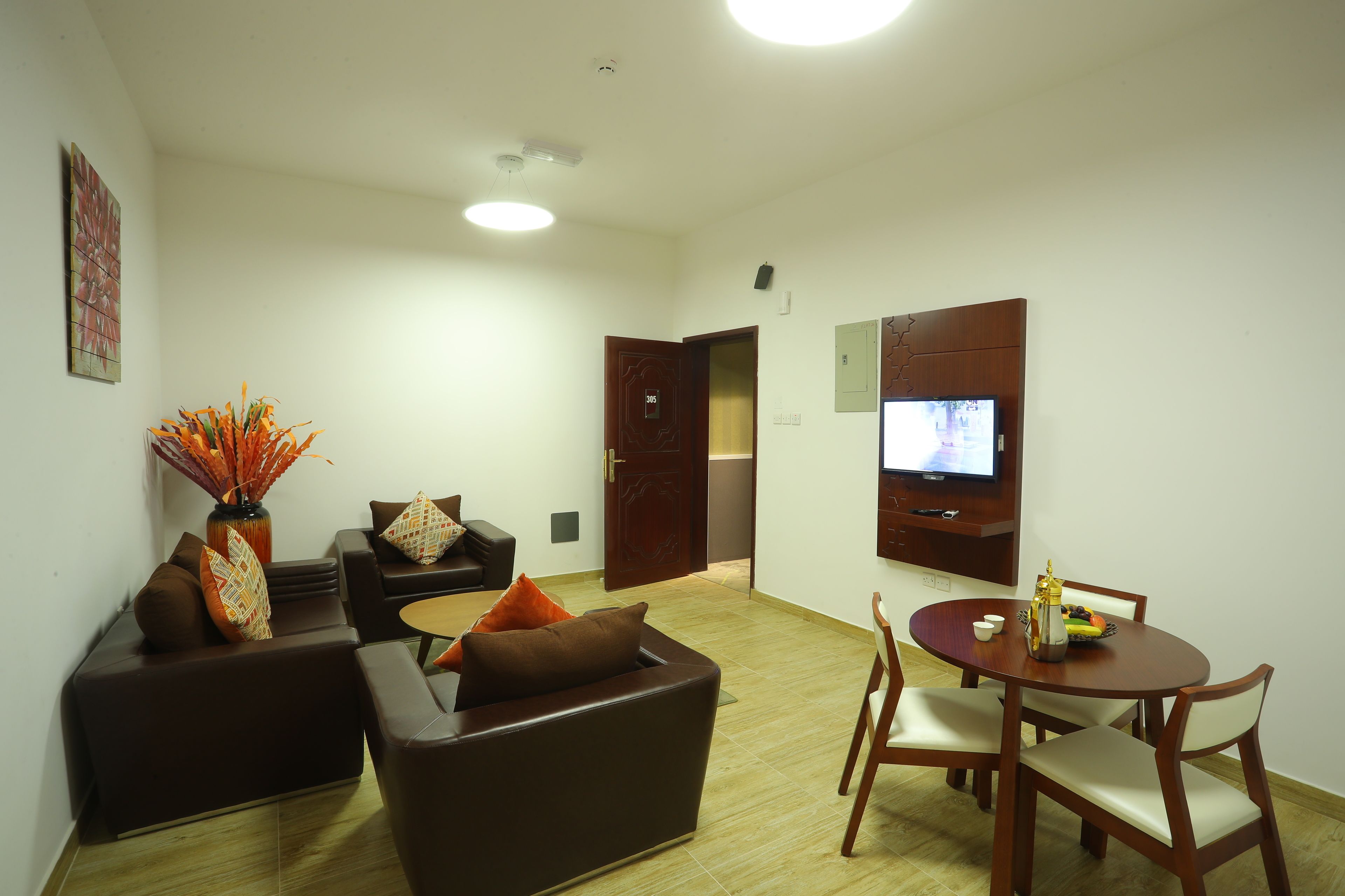 A modern living room with a sofa set, a dining table with chairs, a vase with flowers, and a television mounted on the wall.
