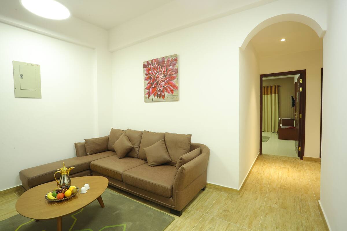 A living room with a brown sofa, a coffee table with fruits and a pitcher, a wall-mounted light fixture, and a door leading to another room.