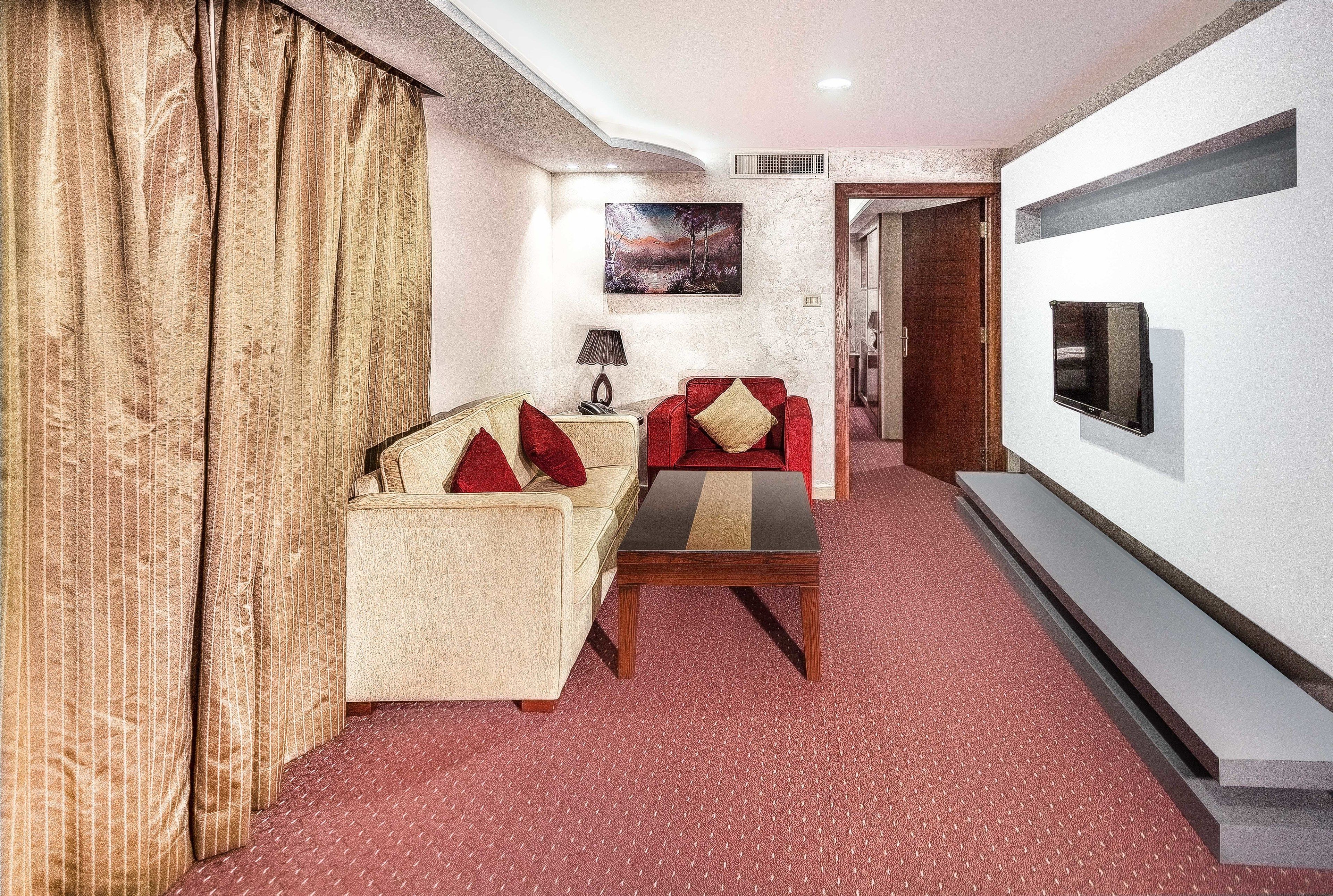 A hotel room with a sofa, armchair, and a small table. There is a painting on the wall and a flat-screen TV mounted on the wall. - Sofia Suites Hotel (Amman)