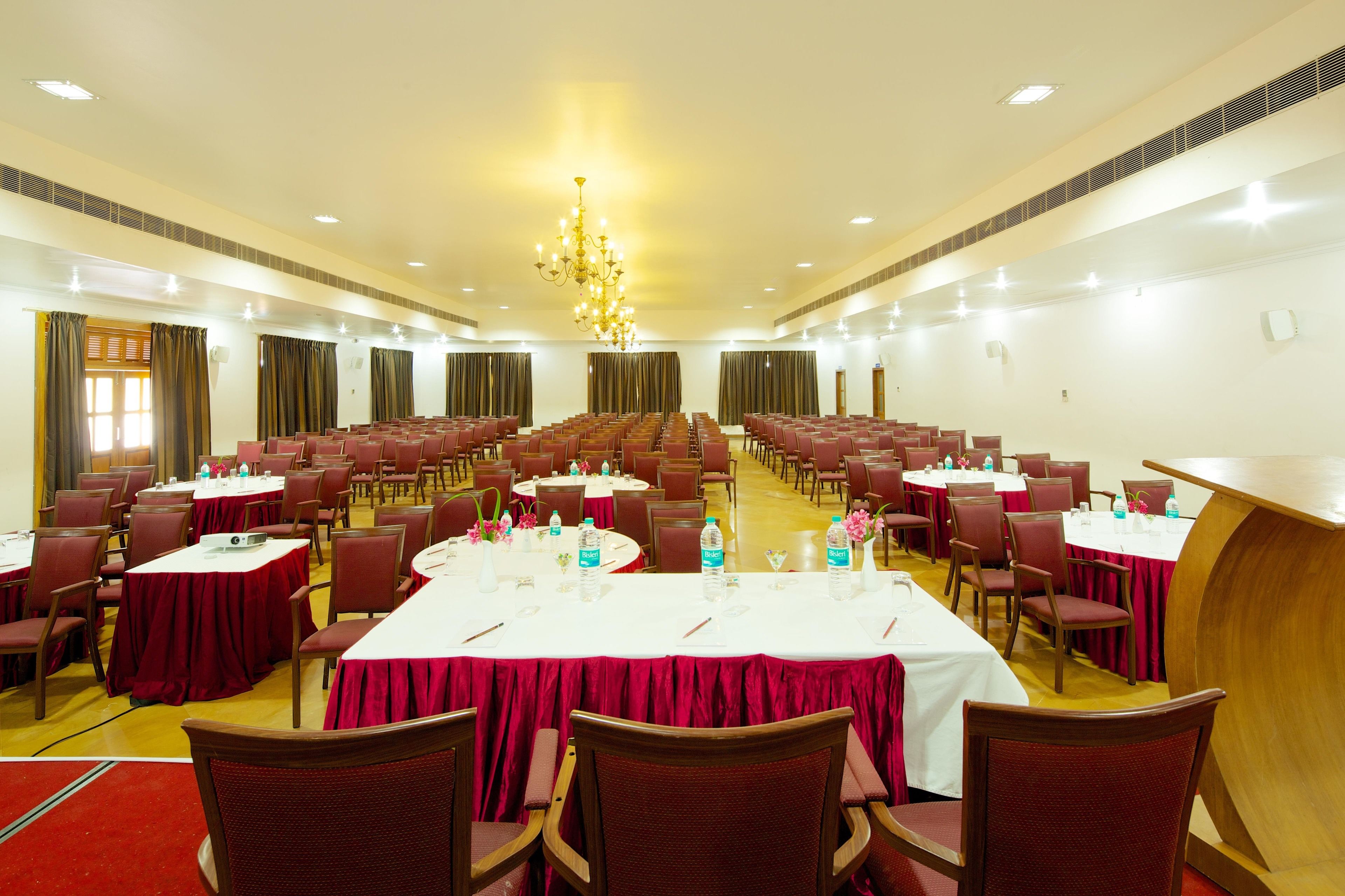 A large conference room with rows of tables and chairs, equipped with water bottles and floral centerpieces.