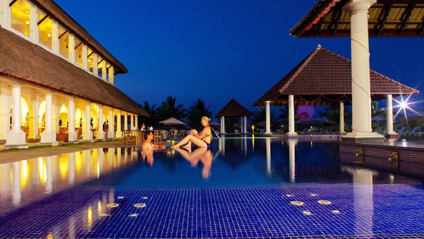 A swimming pool with tiled flooring and a thatched-roof building in the background. - Le Pondy Beach & Lake Resort (Puducherry)