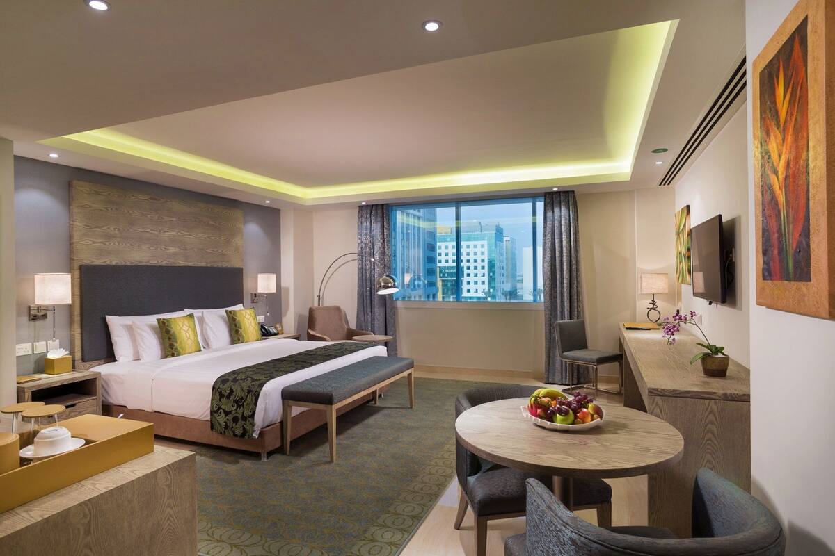 Habitación de hotel con cama, mesita de noche, lámpara, escritorio, silla y una mesa redonda con fruta.